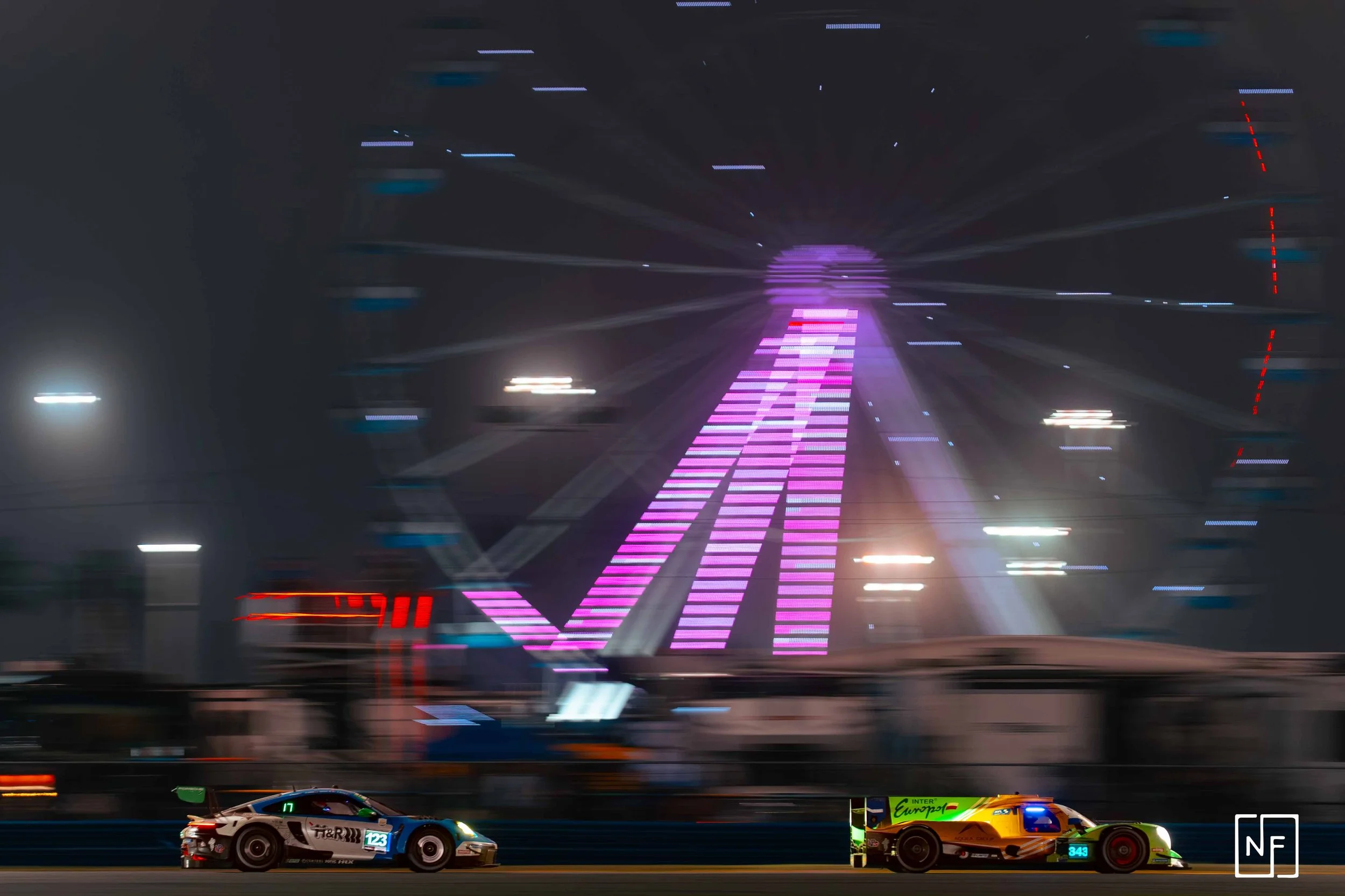 Nighttime scene of a racing track with two race cars in foreground, one black and white with the number 123, the other orange and green with the number 343. In the background, a large illuminated sign displays the word 'FUN' in pink and purple lights