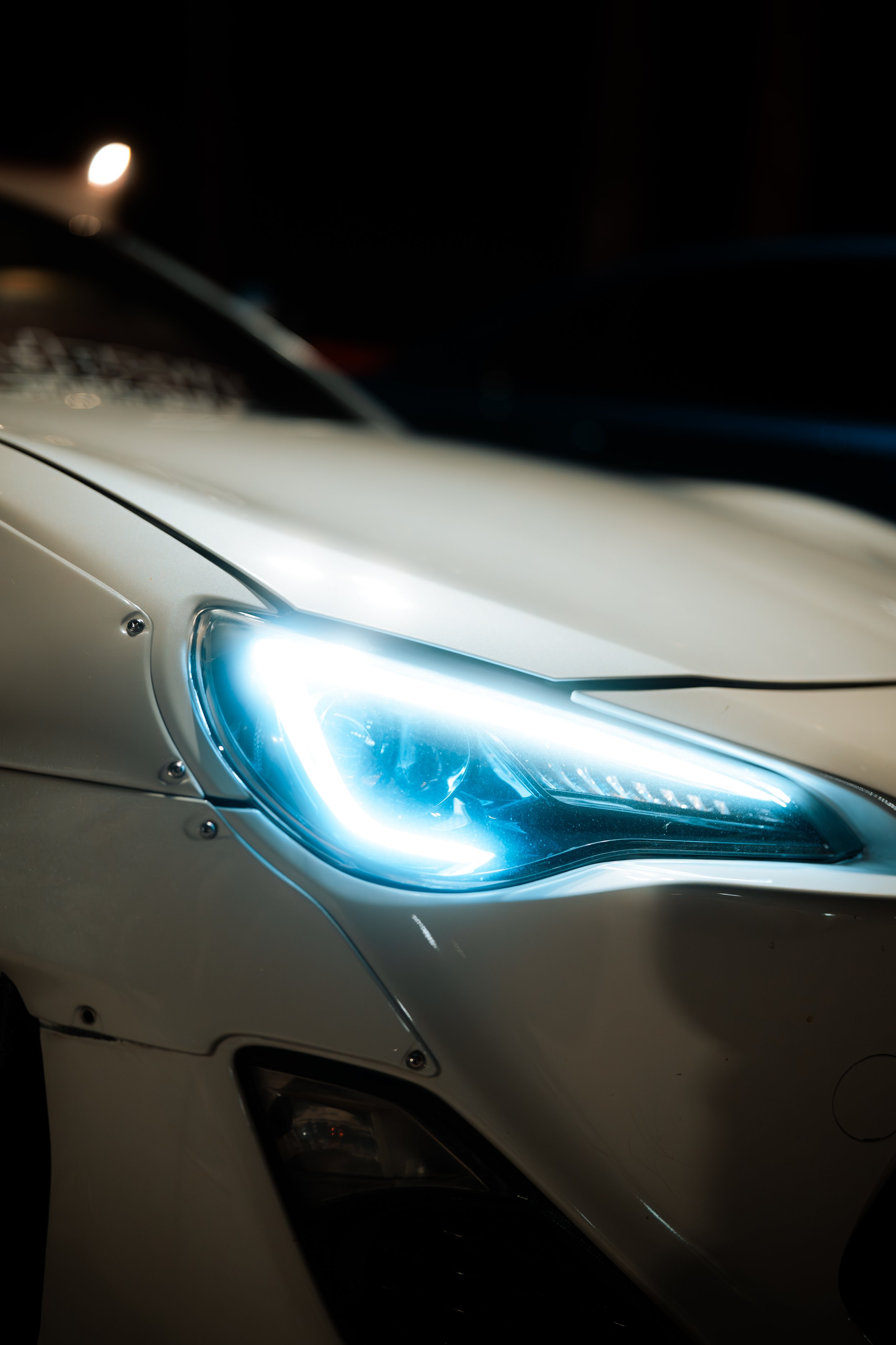 Close-up of a white car's front headlight illuminated at night.