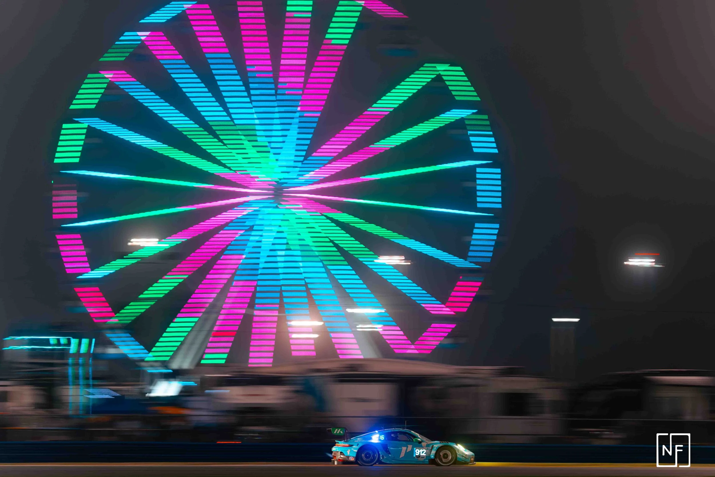 A race car on a dark track in front of a brightly lit Ferris wheel with neon blue, pink, and green lights at night.