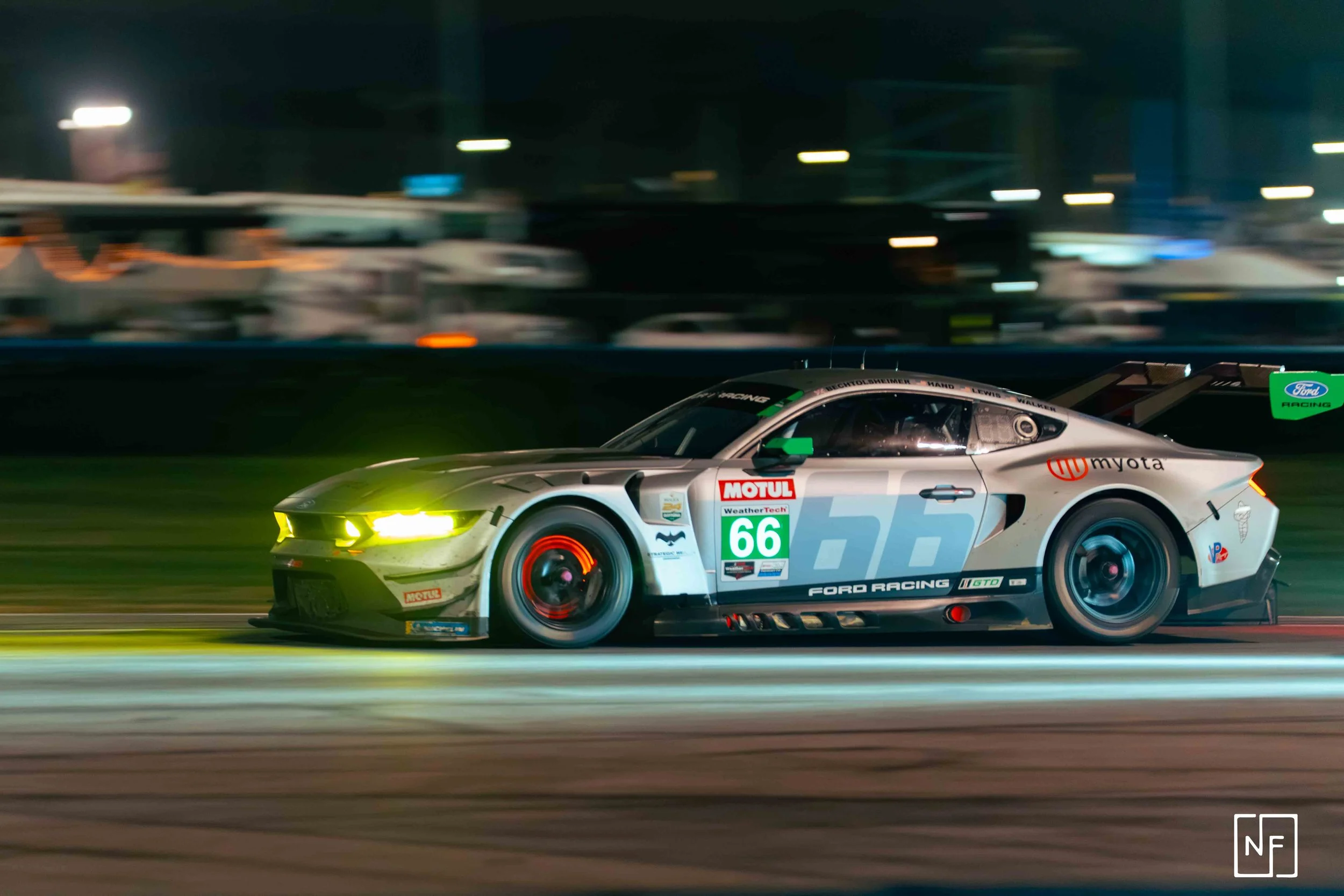 A silver and black Ford race car with bright yellow headlights racing at night, with blurred background.