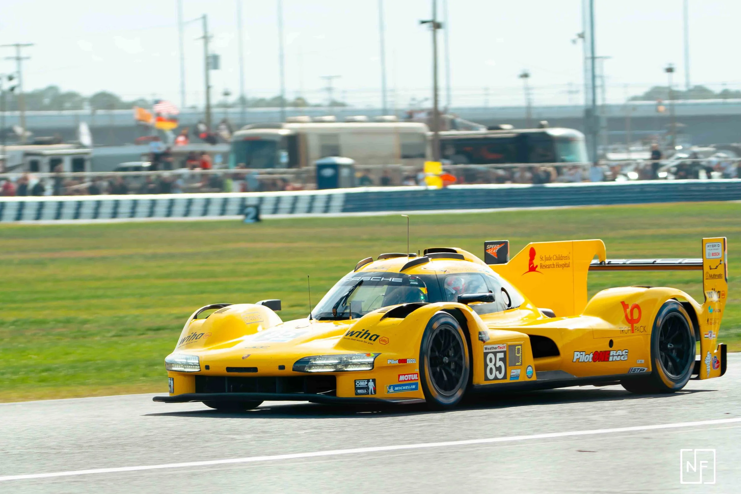 A yellow racing car on a track during a race with a crowd of spectators and vehicles in the background.