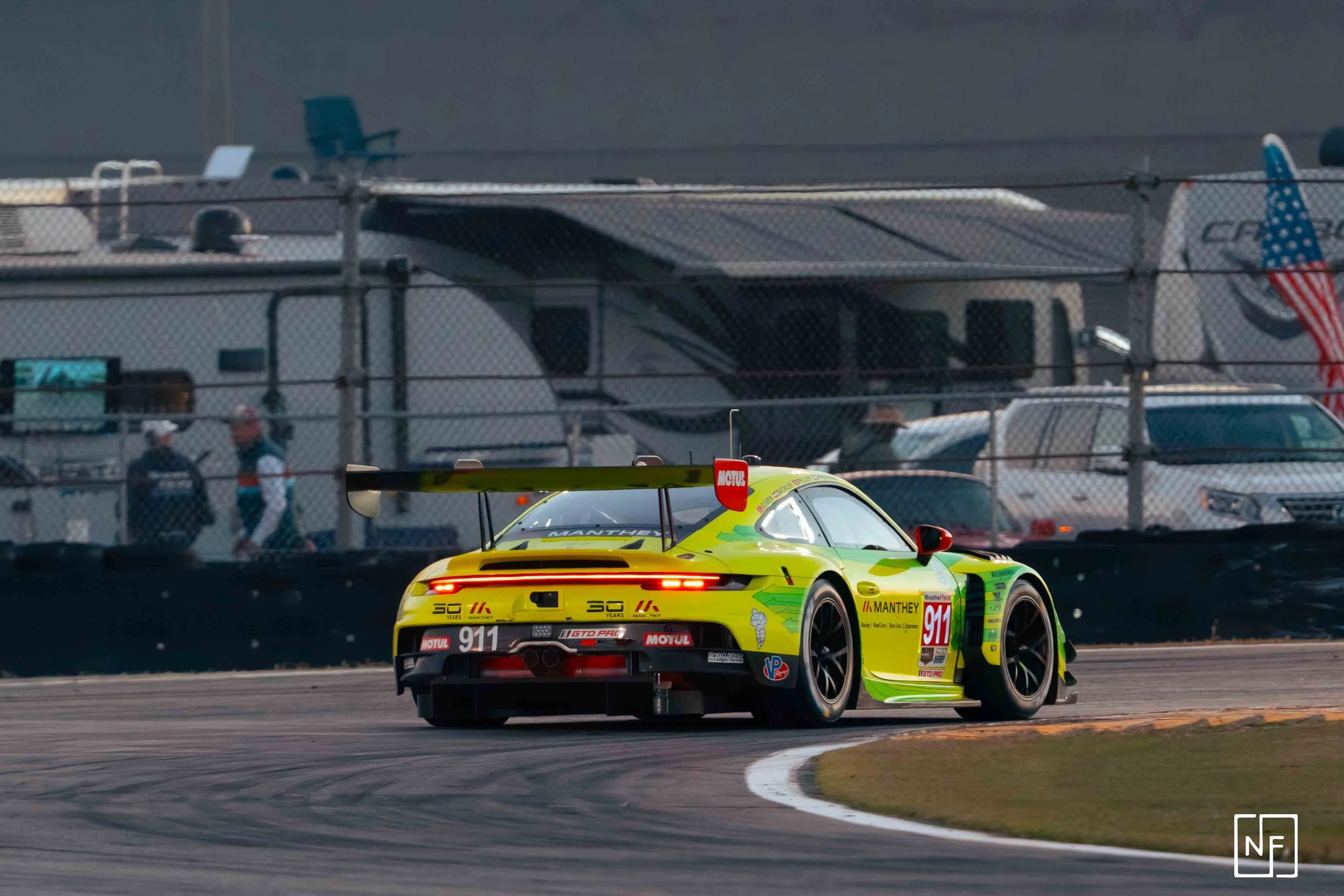 A yellow and green race car on a racetrack, with a large rear wing, close to the curve, and a fence with vehicles and a motorhome in the background.