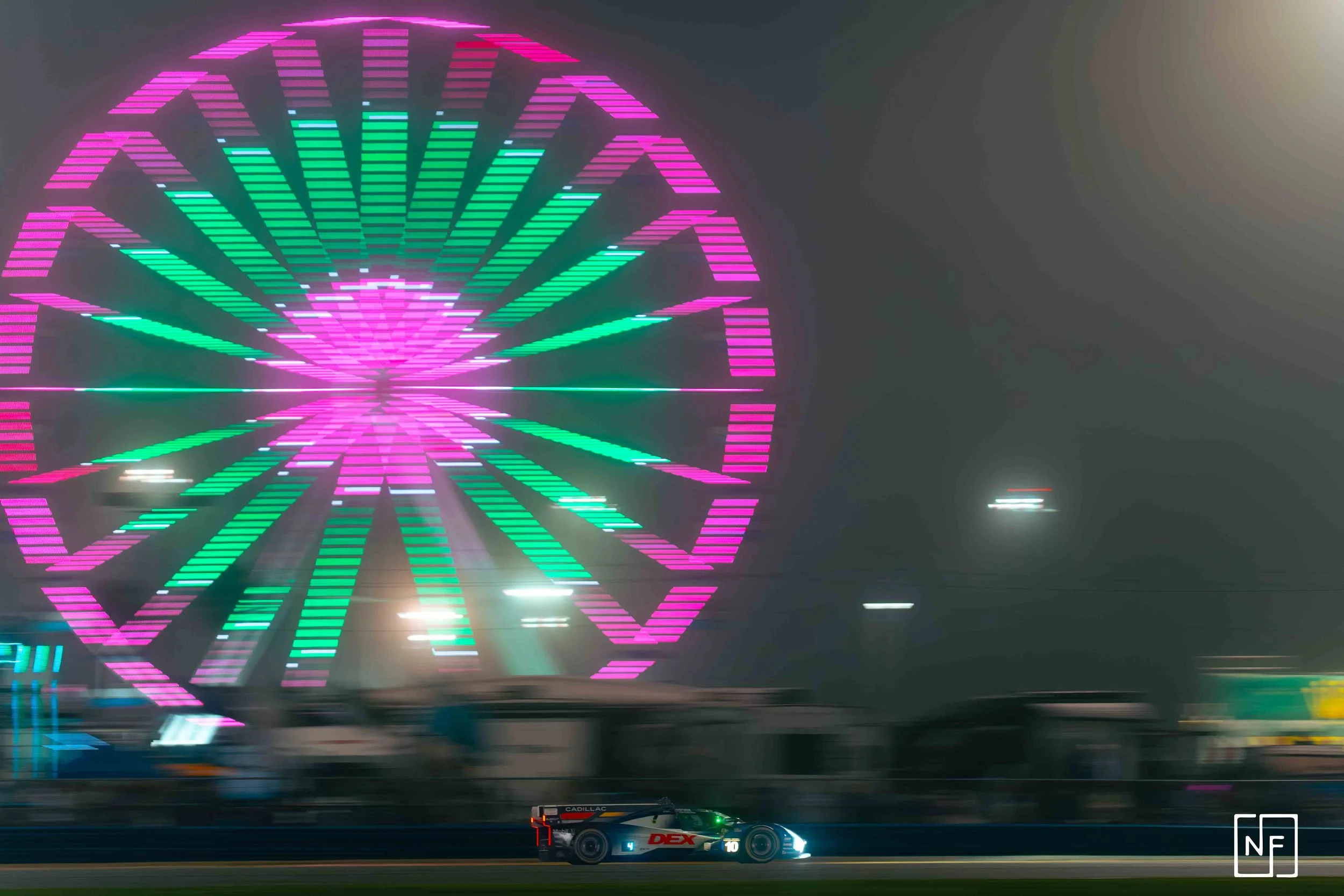 Nighttime scene with a bright pink and green illuminated Ferris wheel in the background and a racing car with branding in the foreground.
