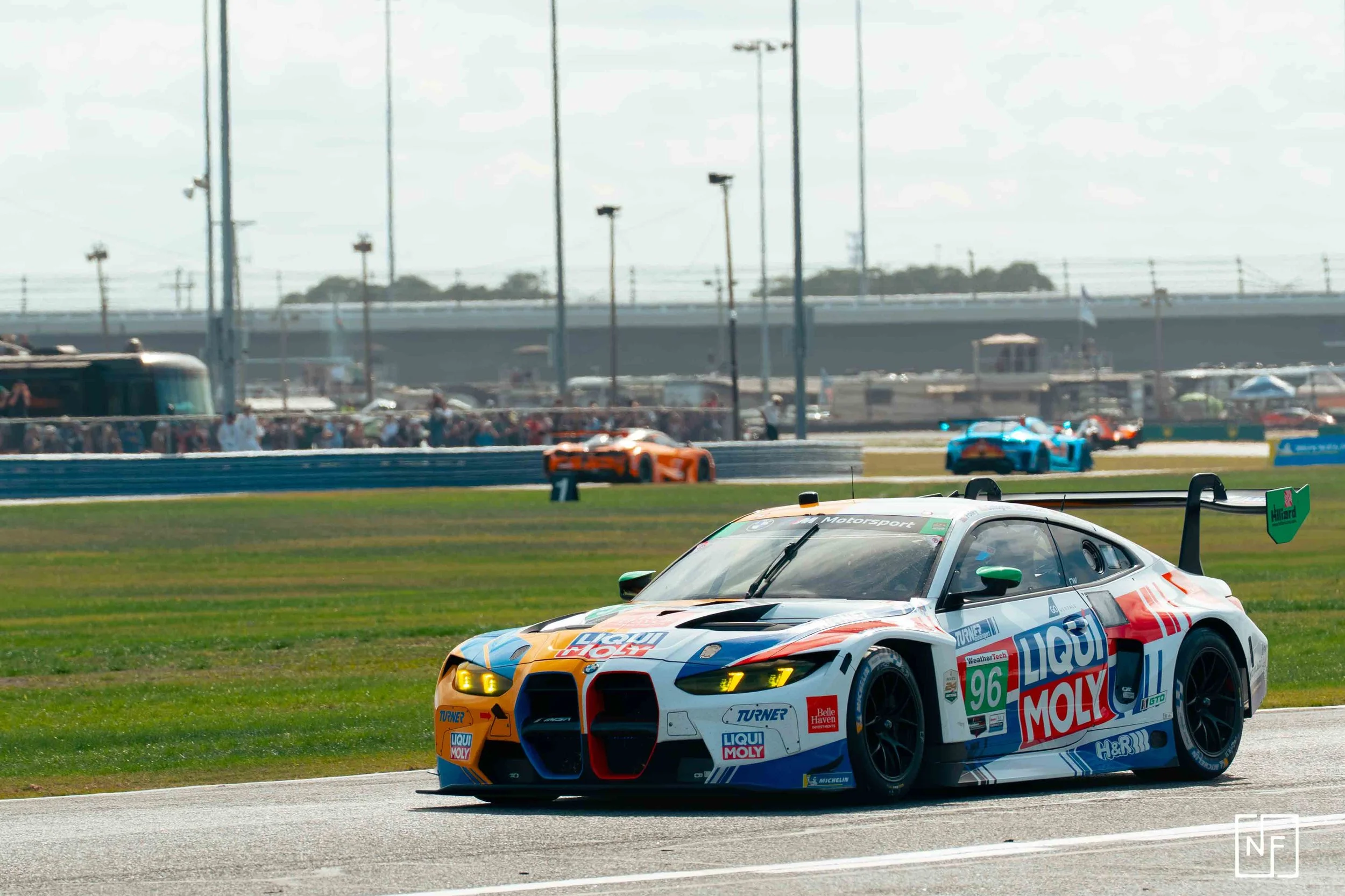 A race car on a racetrack with other race cars in the background and spectators watching from the stands.
