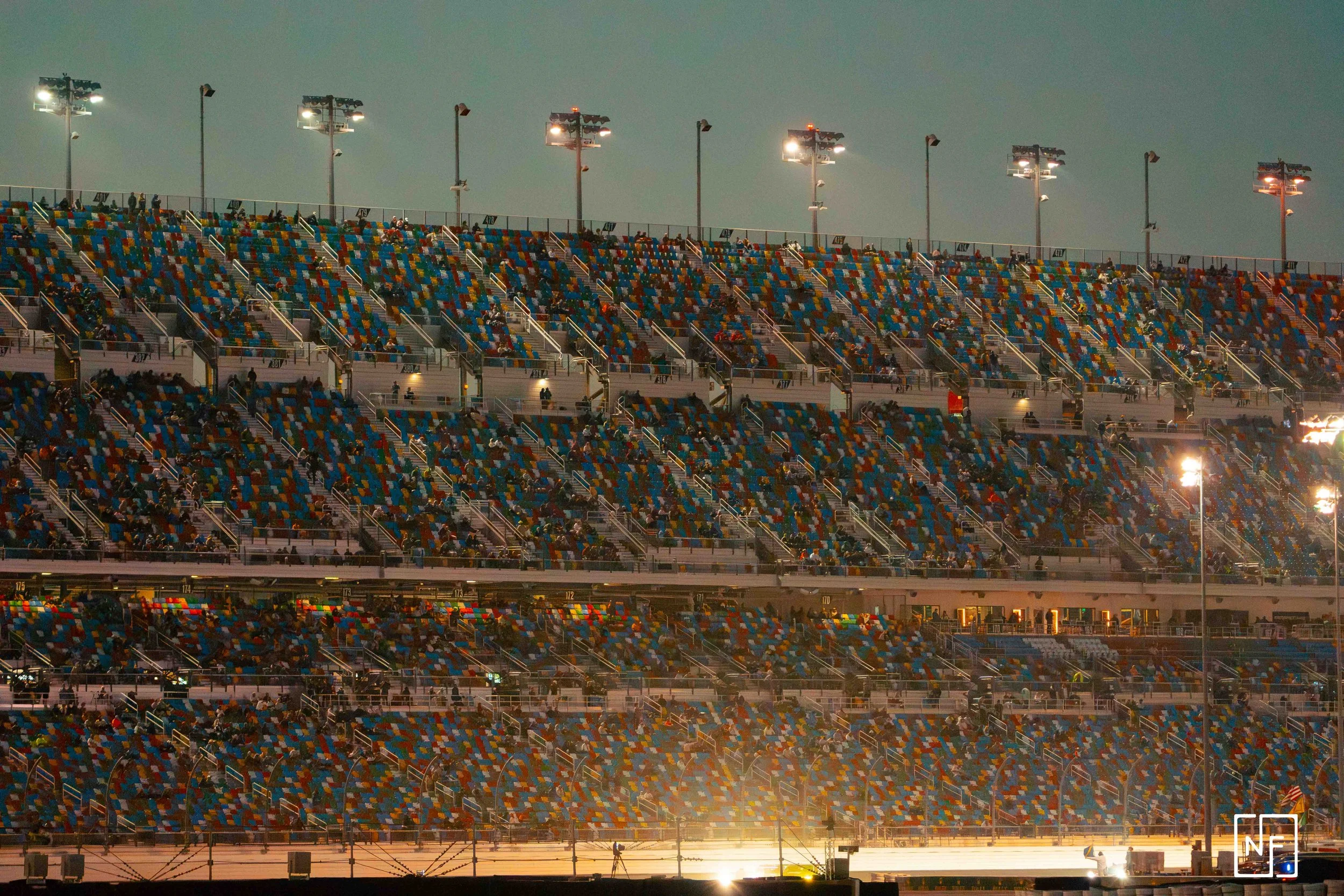 Empty multi-level grandstand at a race track illuminated by bright lights during dusk, with a few spectators seated.