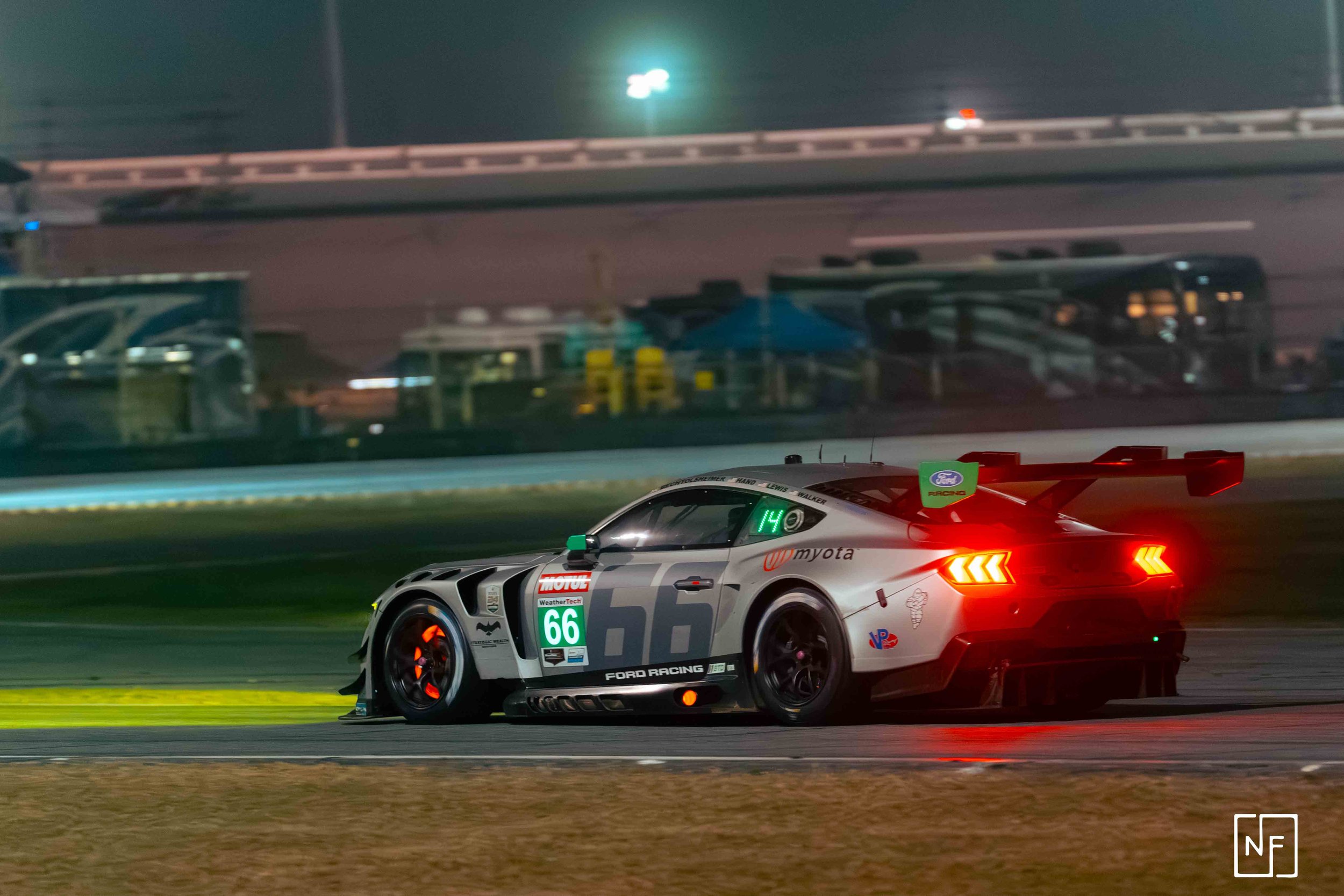 A Ford racing car with the number 66 on its side drives on a race track at night, illuminated by red tail lights and surrounded by blurred background.