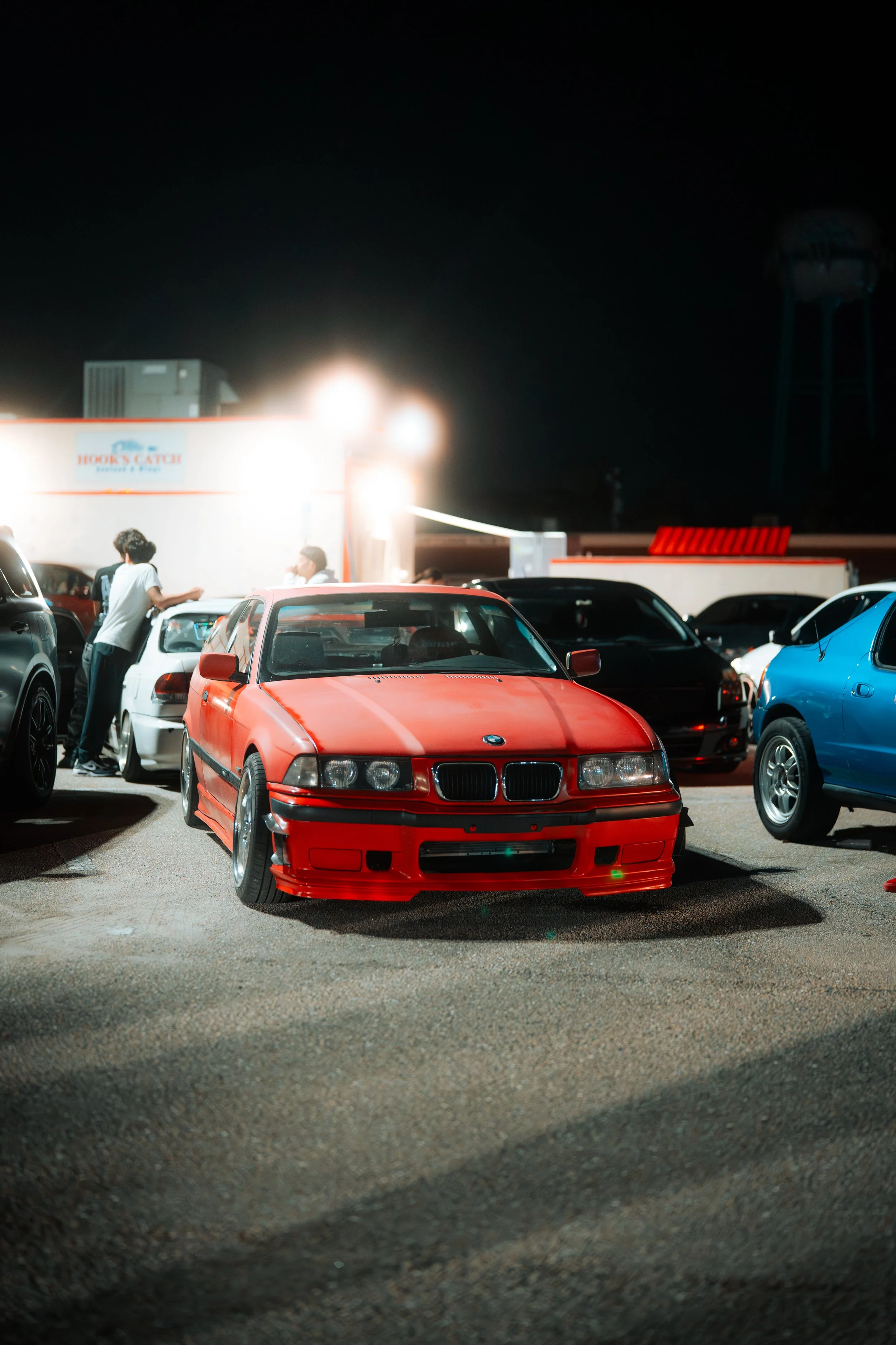 A red BMW parked in a lot at night, surrounded by other cars and a few people conversing in the background.