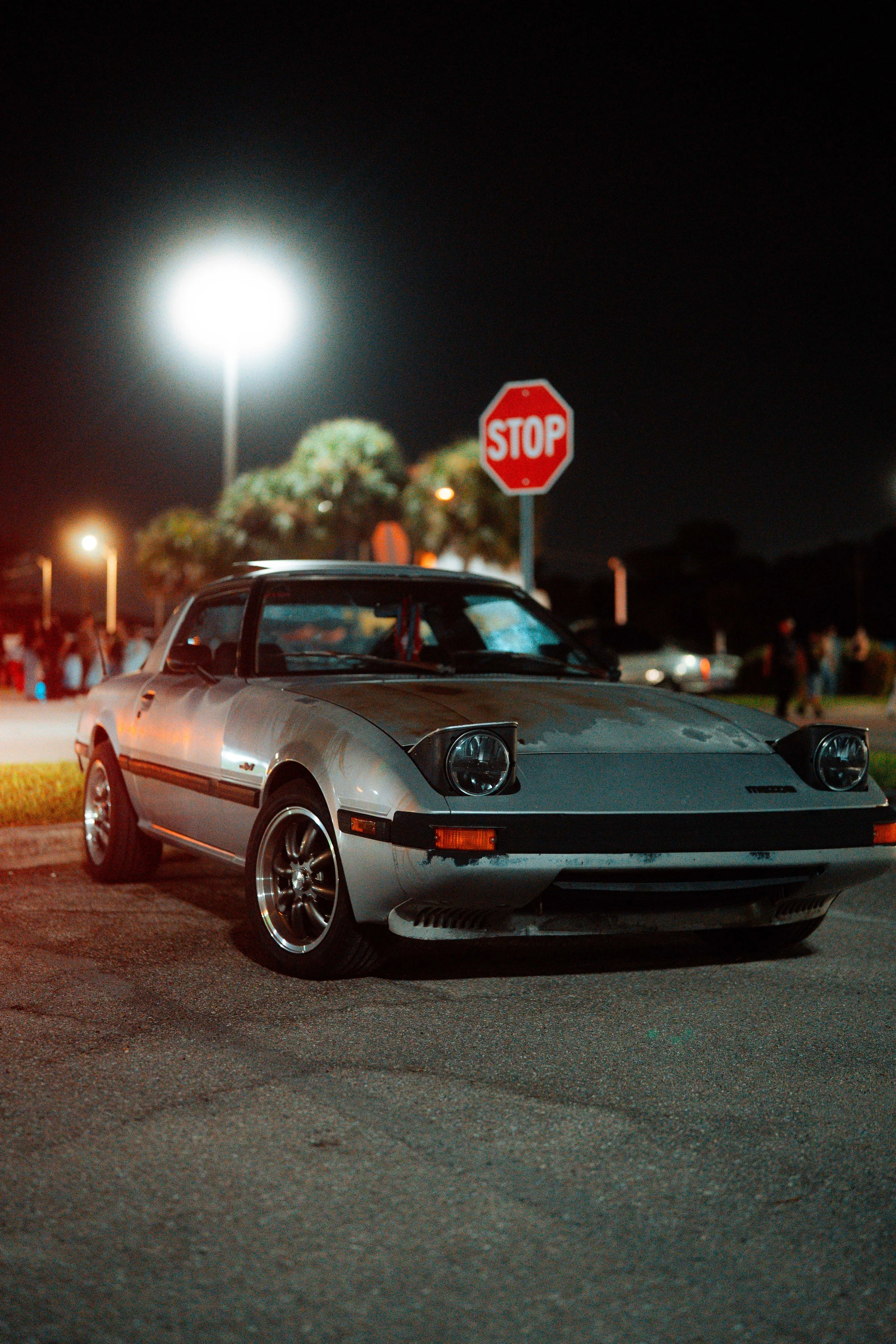 A vintage silver sports car parked at night near a stop sign in a parking lot with people and trees in the background.