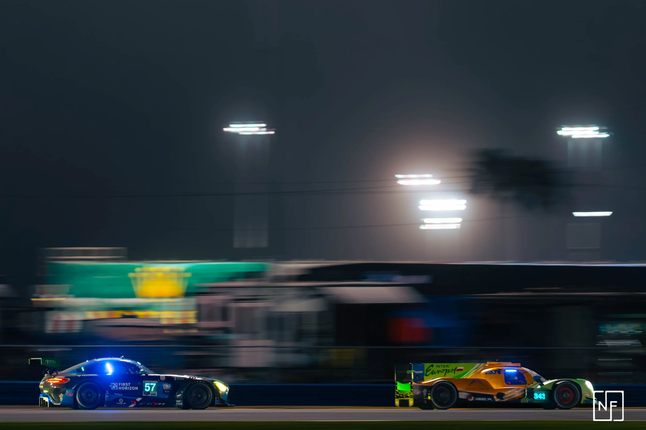 Nighttime racing scene featuring two race cars on a track, one blue with the number 57 and one orange with the number 343, blurring motion effects with bright lights in the background.
