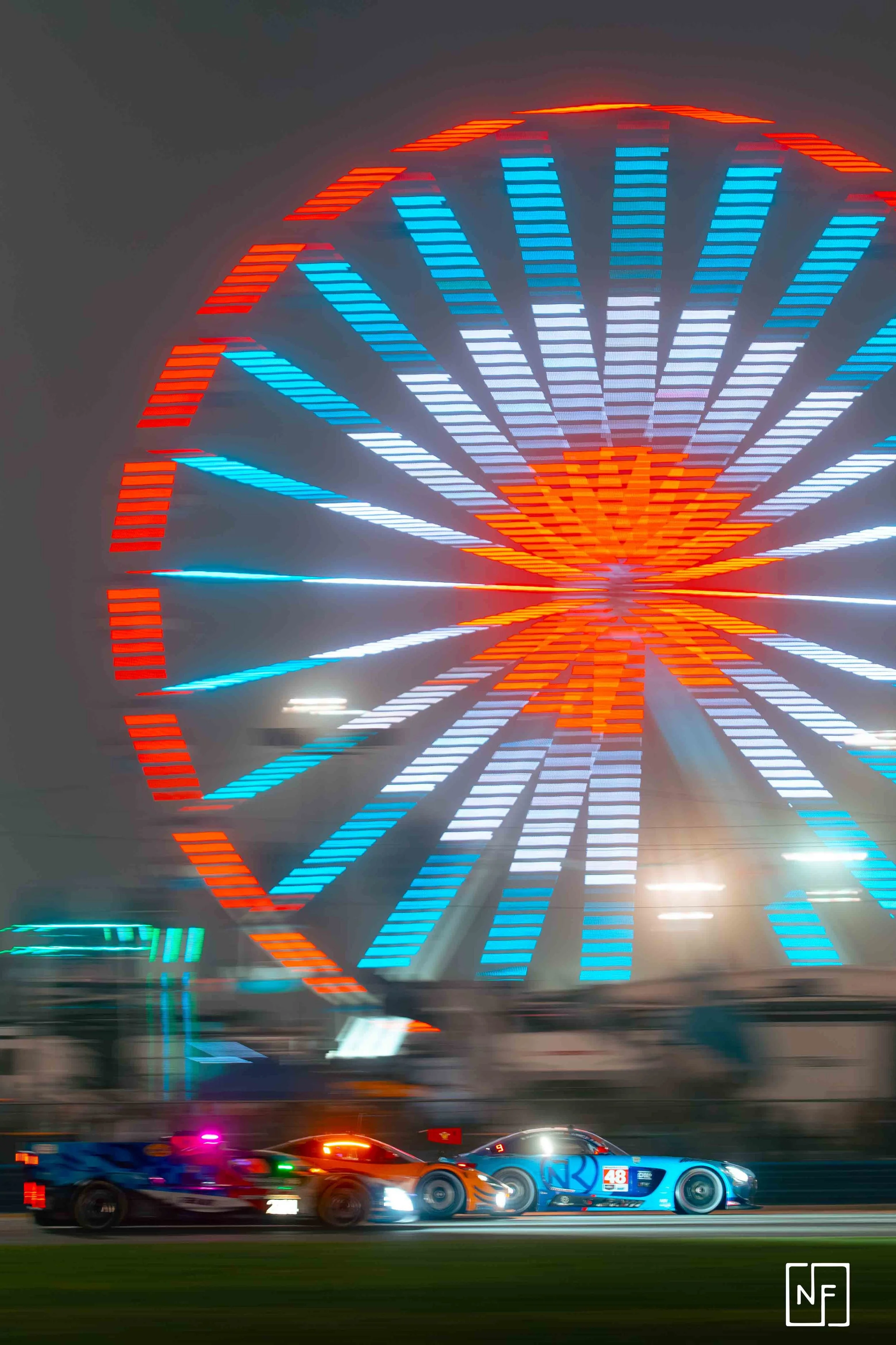 Nighttime photo of a brightly lit Ferris wheel with red, blue, and white lights in motion, with race cars on the track in the foreground, creating a blurred effect.
