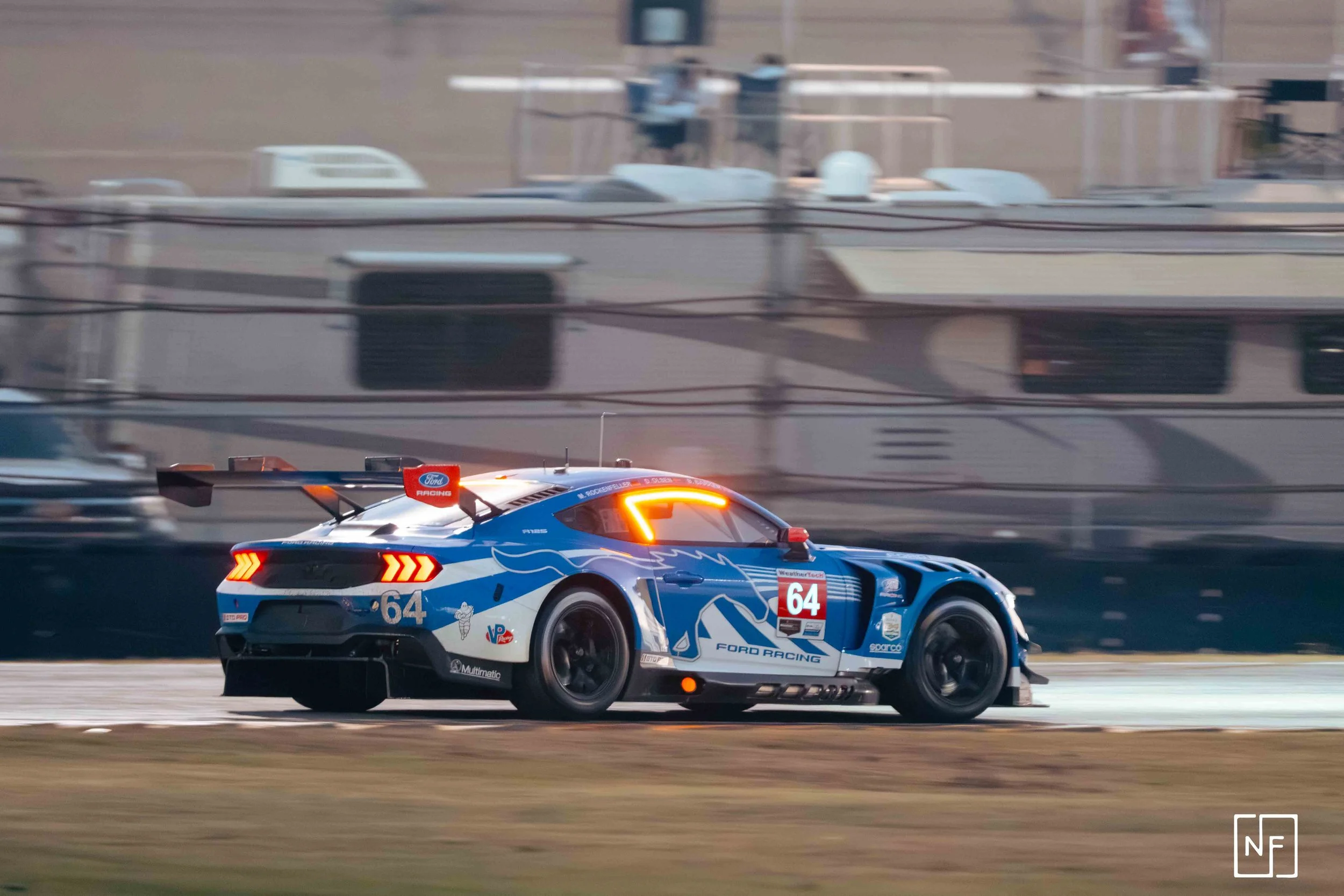 A blue and white Ford racing car with the number 64 on its side, speeding on a race track, with blurred background of trailers and equipment.