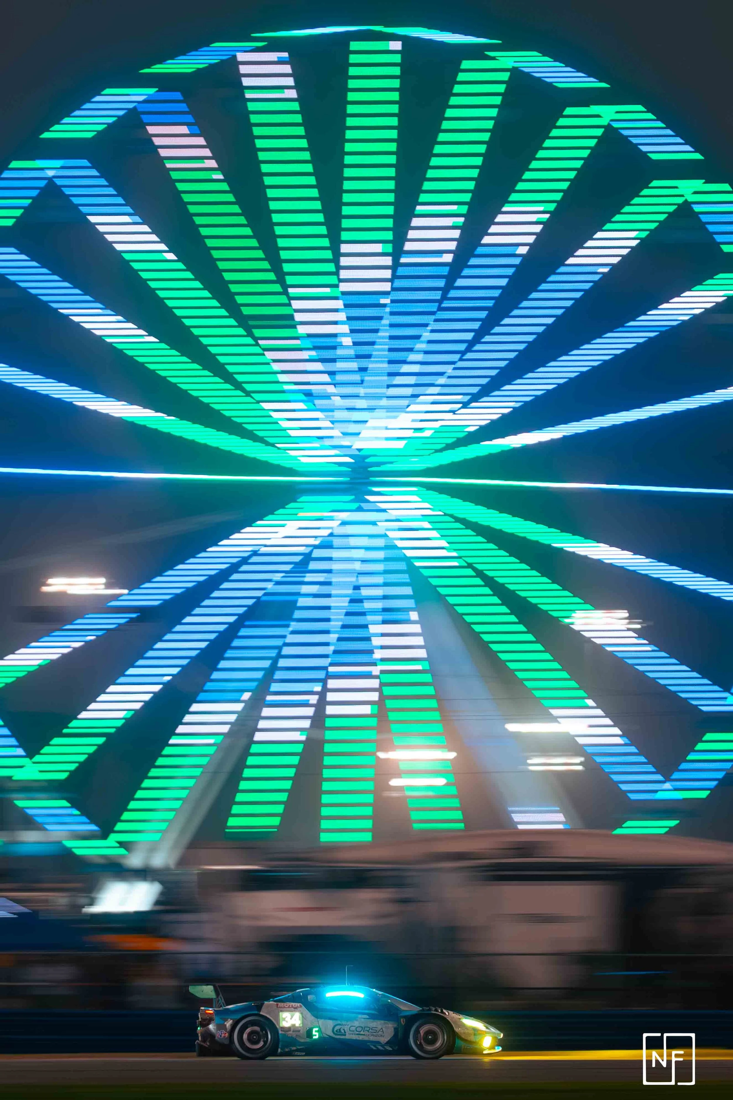 Nighttime scene of a race car passing in front of an illuminated Ferris wheel with colorful lights.