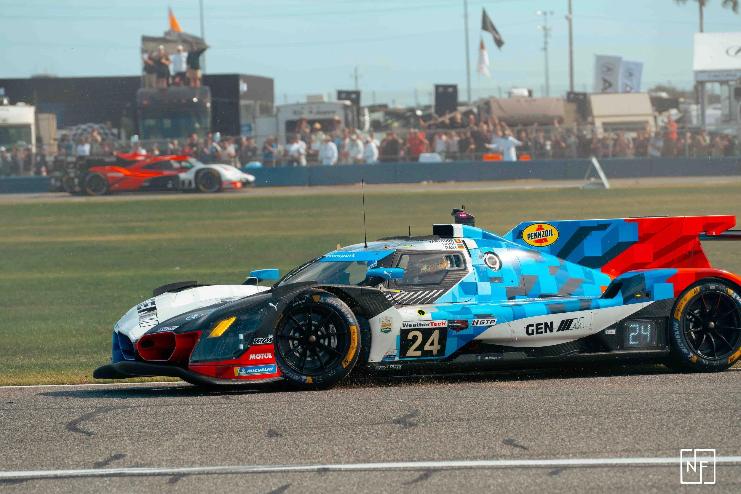 A race car speeding on a track, with a group of cars and spectators in the background.