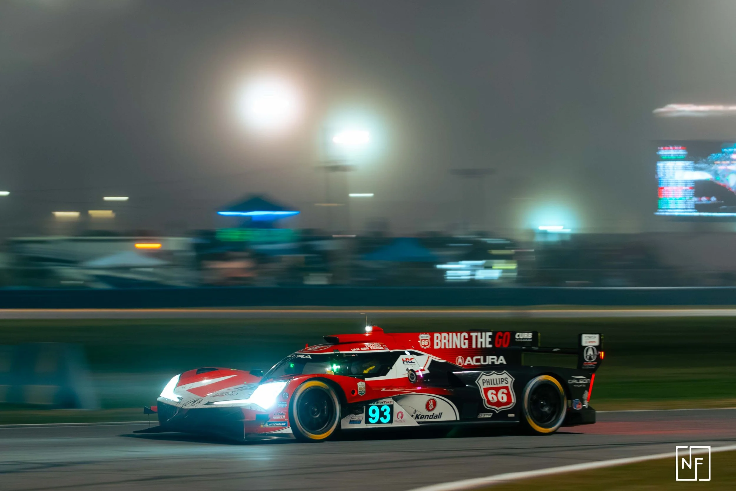 A race car driving on a track at night with headlights on, sponsorship logos, and a blurred background of tents and digital screens.