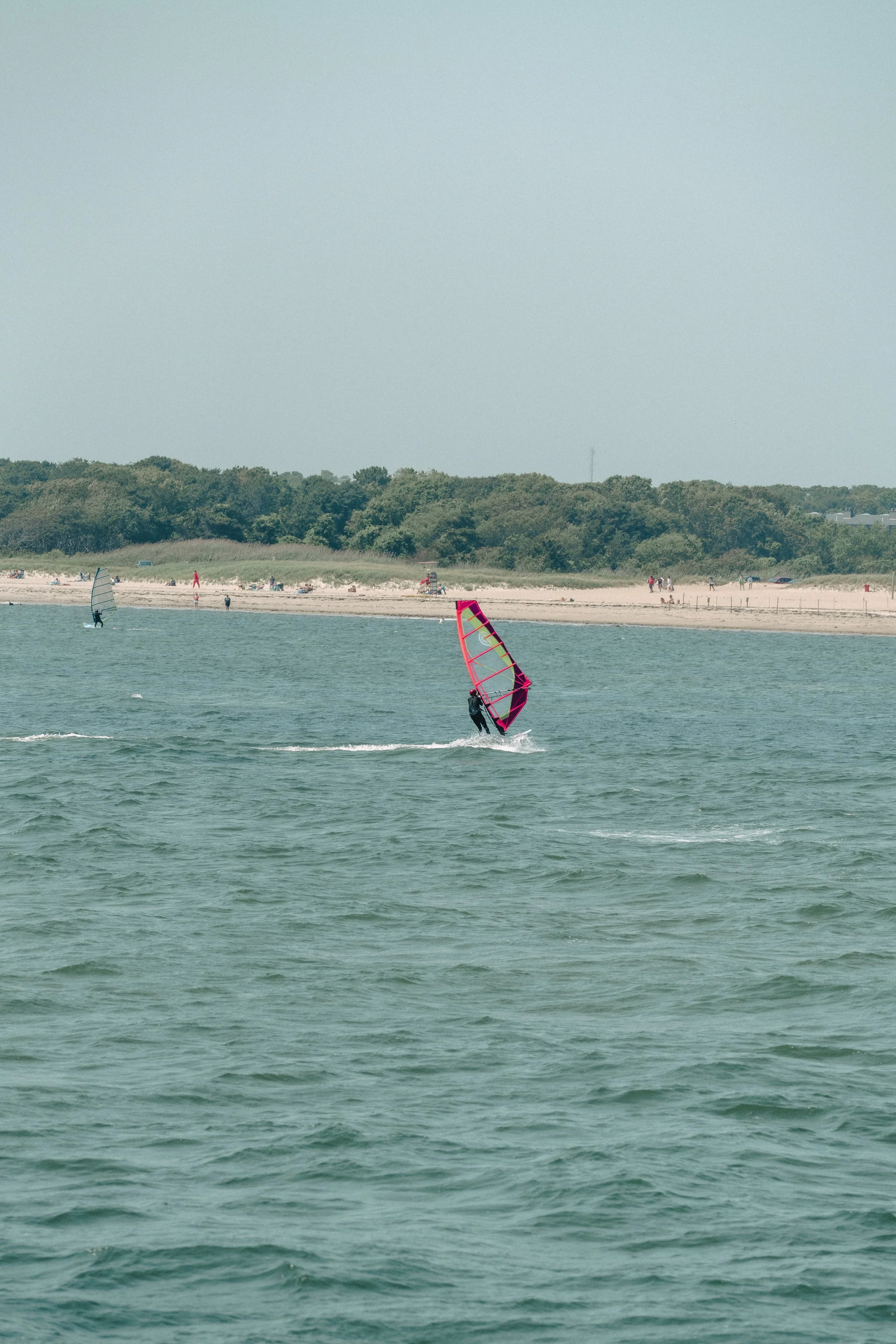 A person windsurfing on the water at the beach with a pink sail, with other windsurfers and people on the beach in the background.