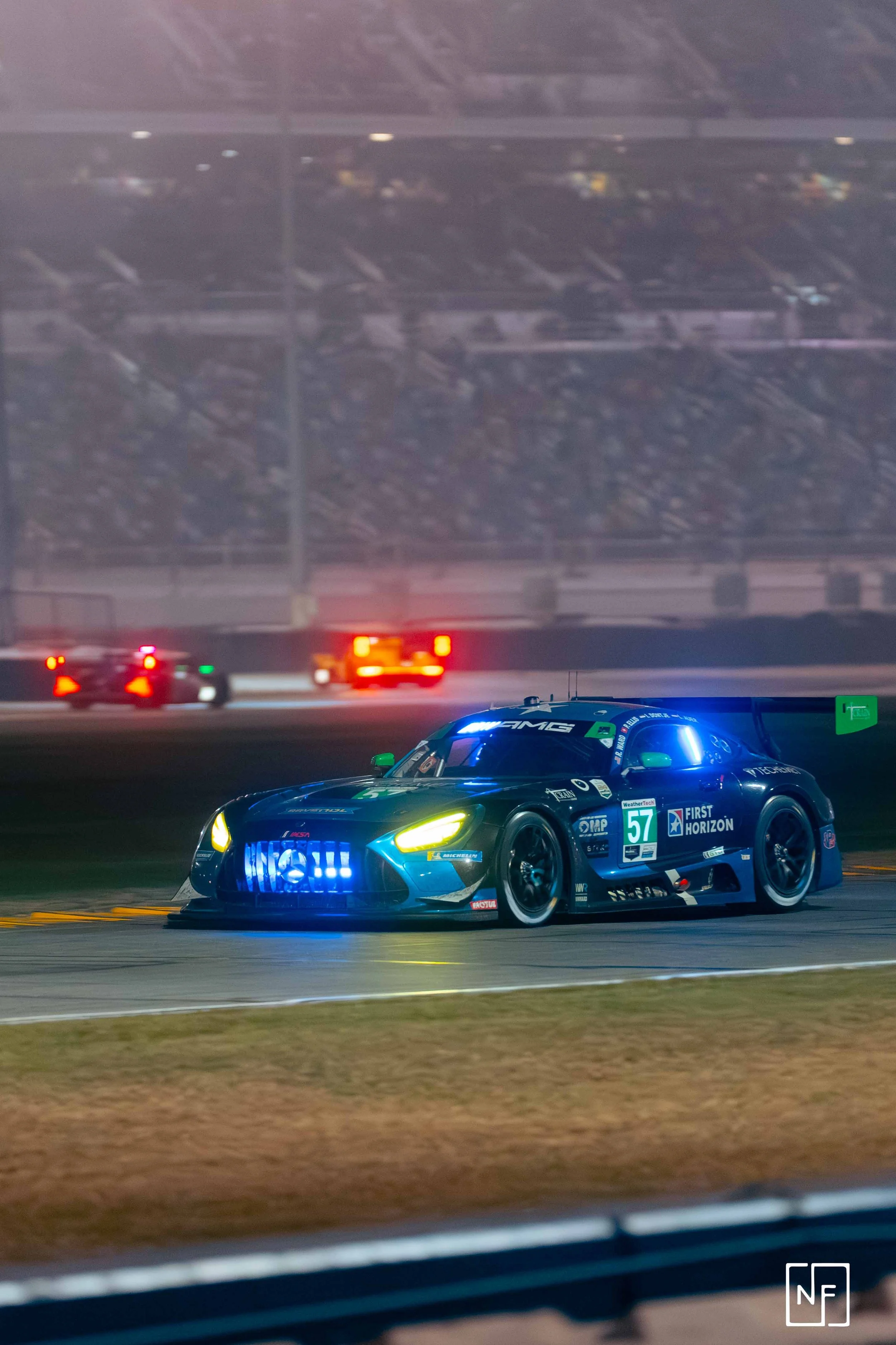 A race car on a track at night, illuminated with blue and yellow lights, with a blurred background and several emergency vehicles in the distance.