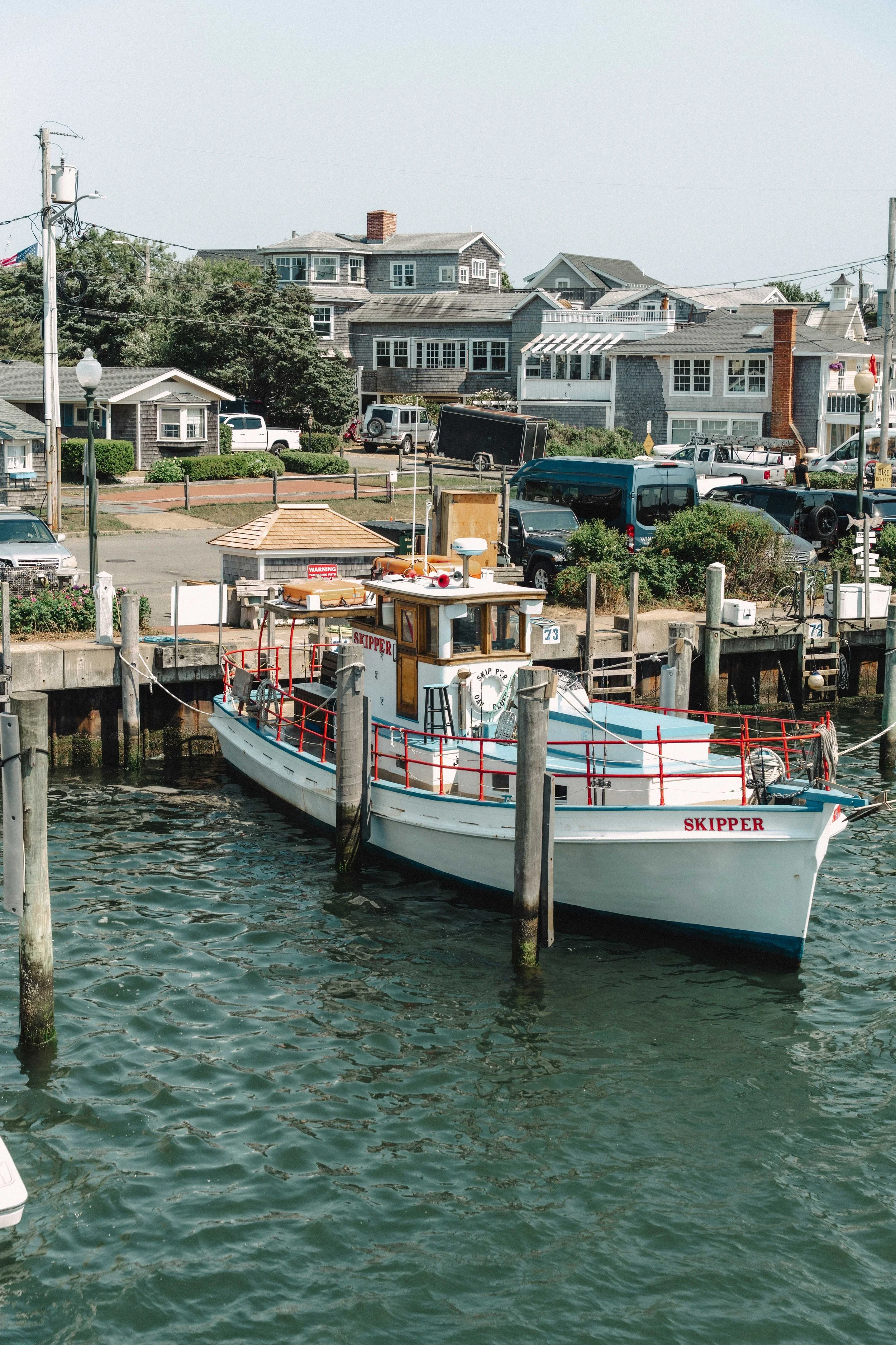A small boat named 'Skipper' docked at a marina with residential houses and parked cars in the background.