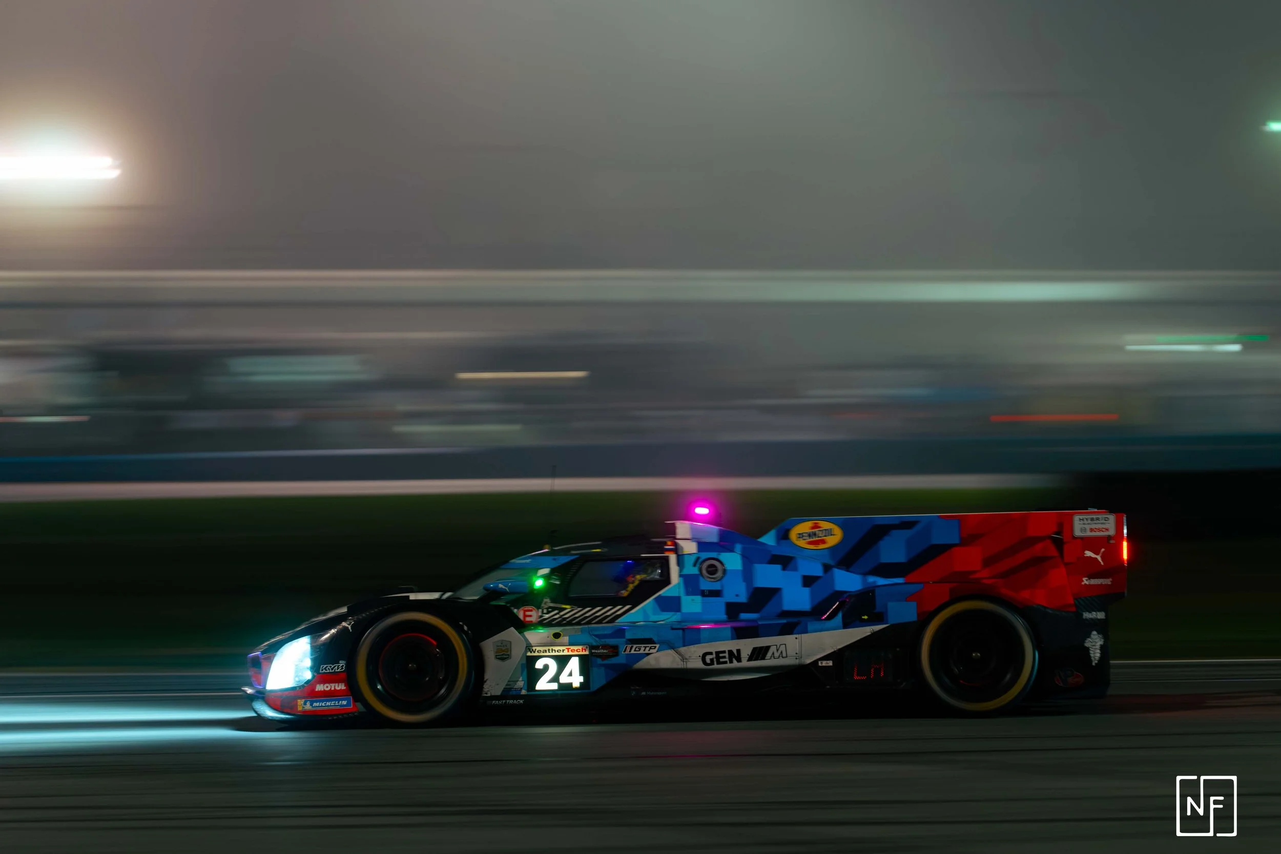 A race car with the number 24 on it speeds on a racetrack at night, with blurred lights and background indicating high speed.