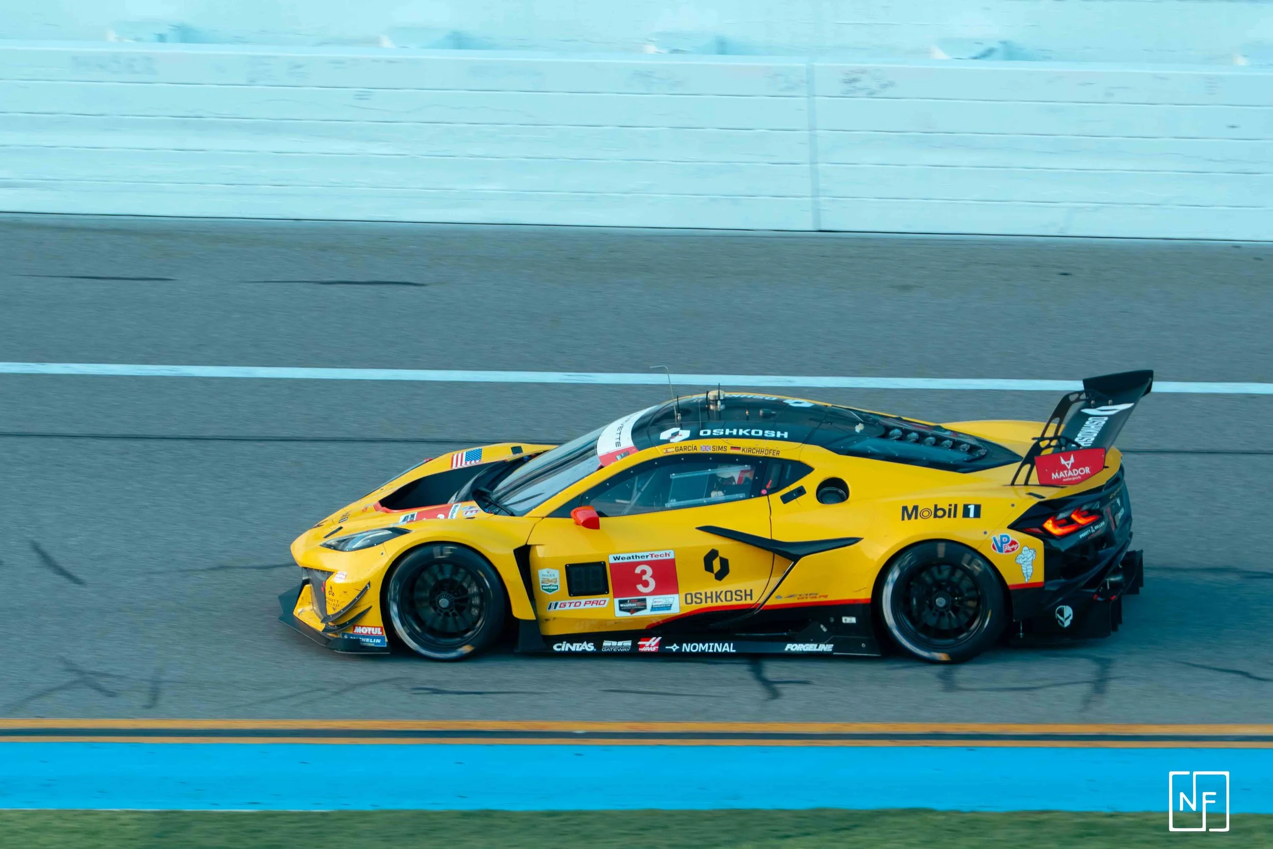 A yellow race car driving on a racetrack, featuring sponsor logos including OshKosh, Mobil 1, and others, with a rear wing and aerodynamic design.