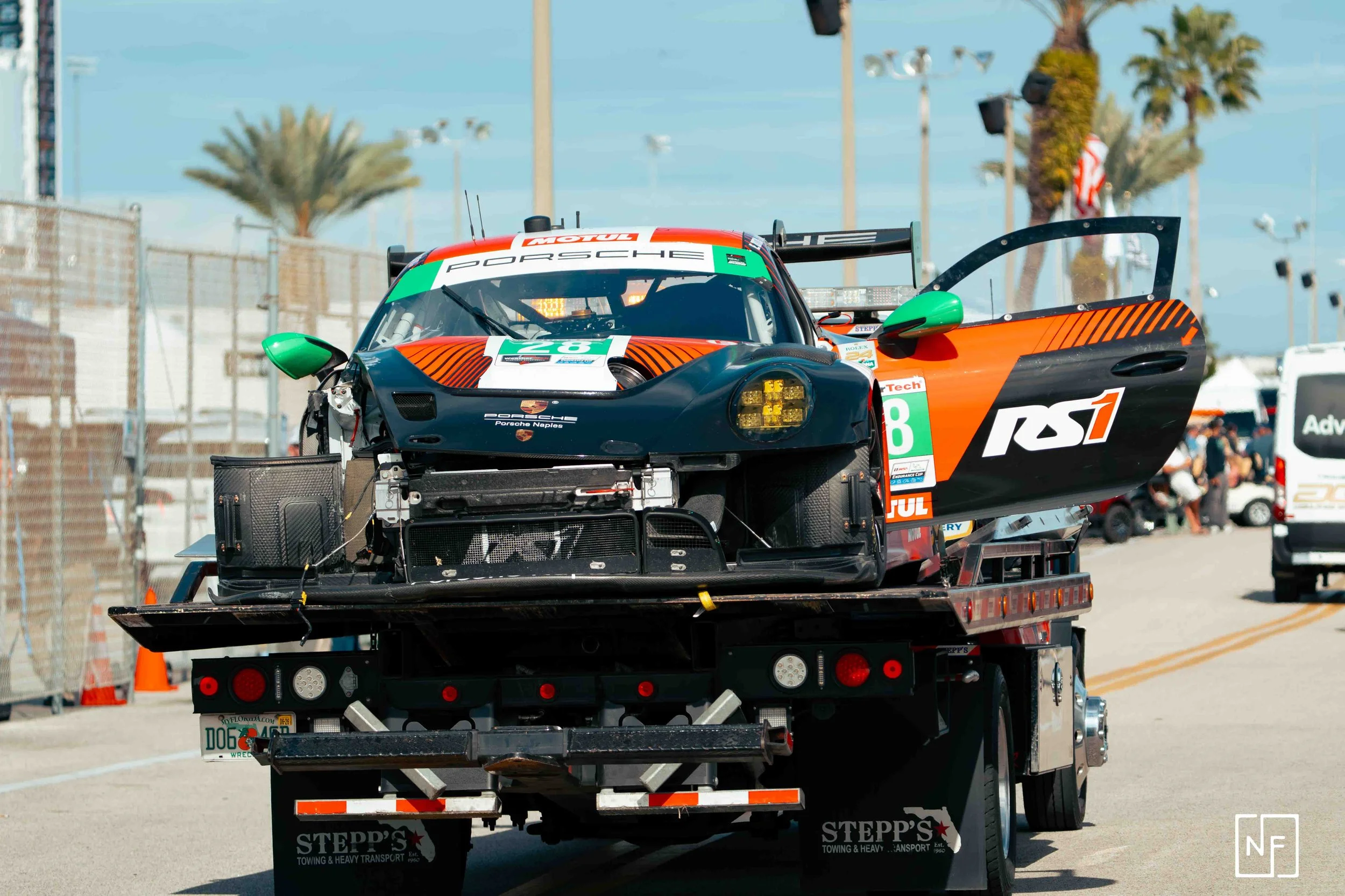 A race car on a tow truck, with the truck's trailer extended. The race car is black, orange, and white, with a partially open door and visible racing decals, including the number 28 and RST logo. The background shows palm trees, a fence, and several 