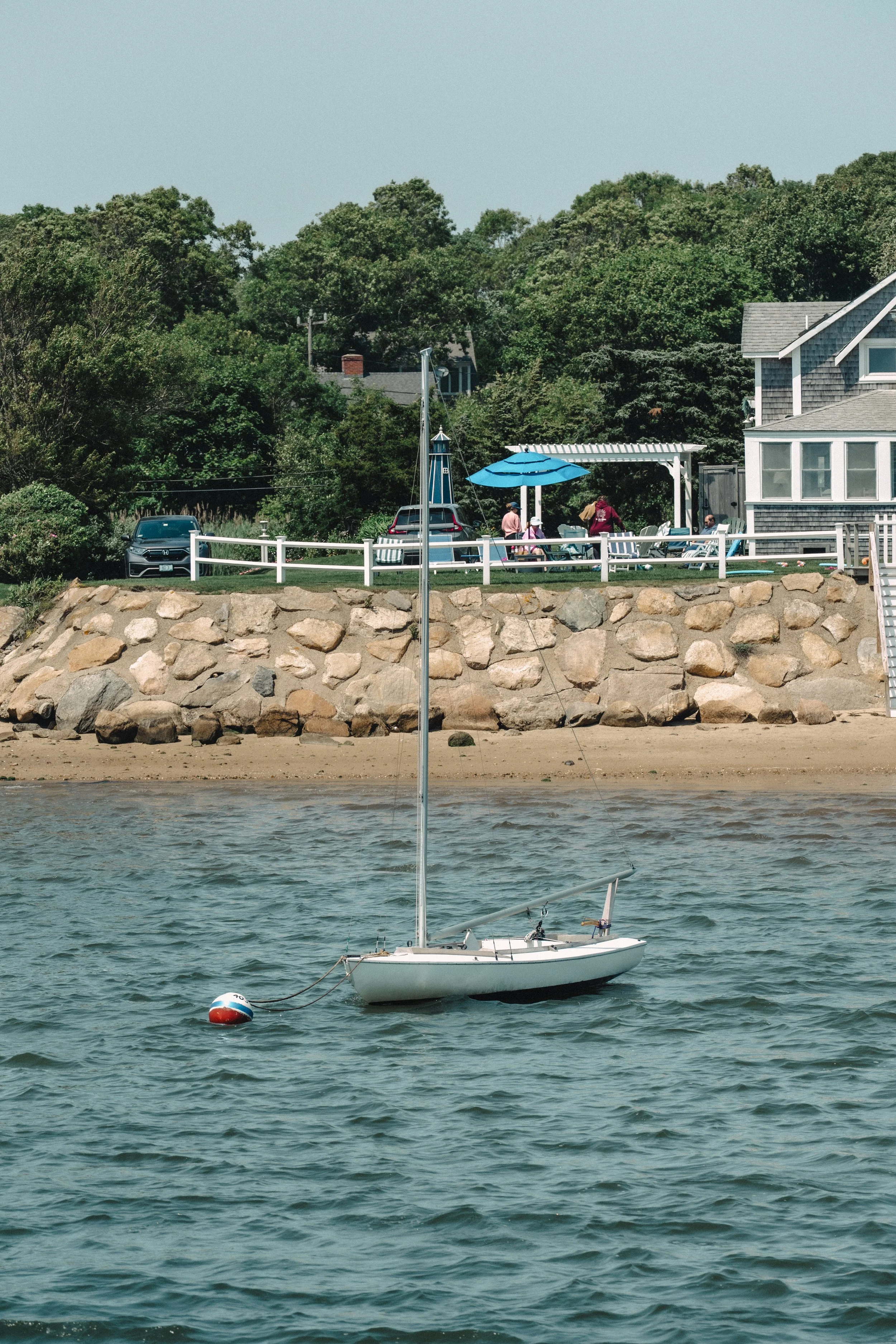 A small sailboat floating on the water near a stone embankment with a house, trees, and people on the shore.