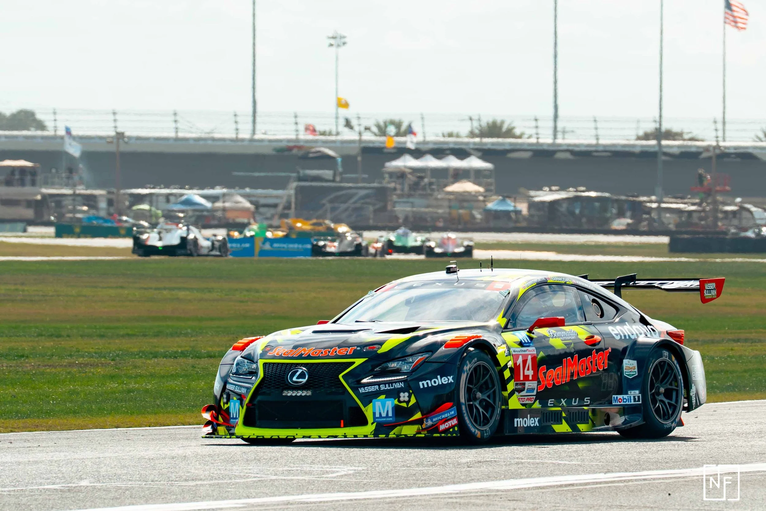 A Lexus race car with sponsors and number 14 on a racetrack during a race, with other racing cars and pit area in the background.