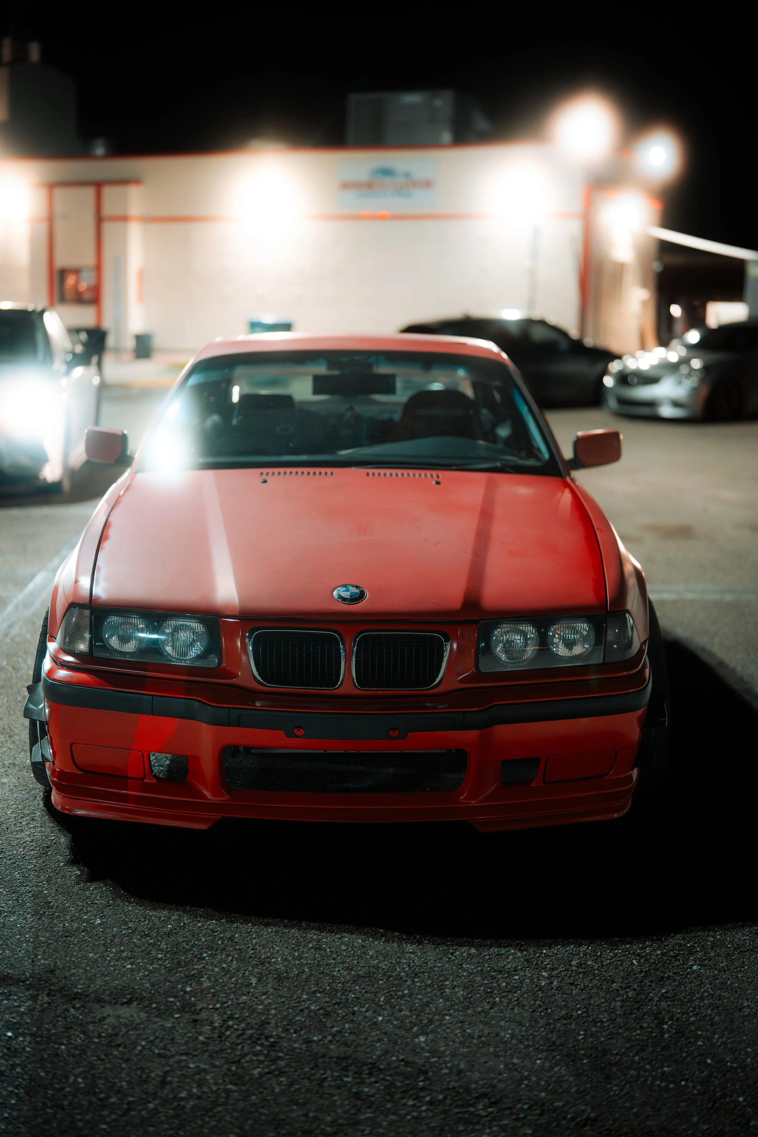 Red BMW parked in a lot at night with blurred background of other cars and a building with bright lights.