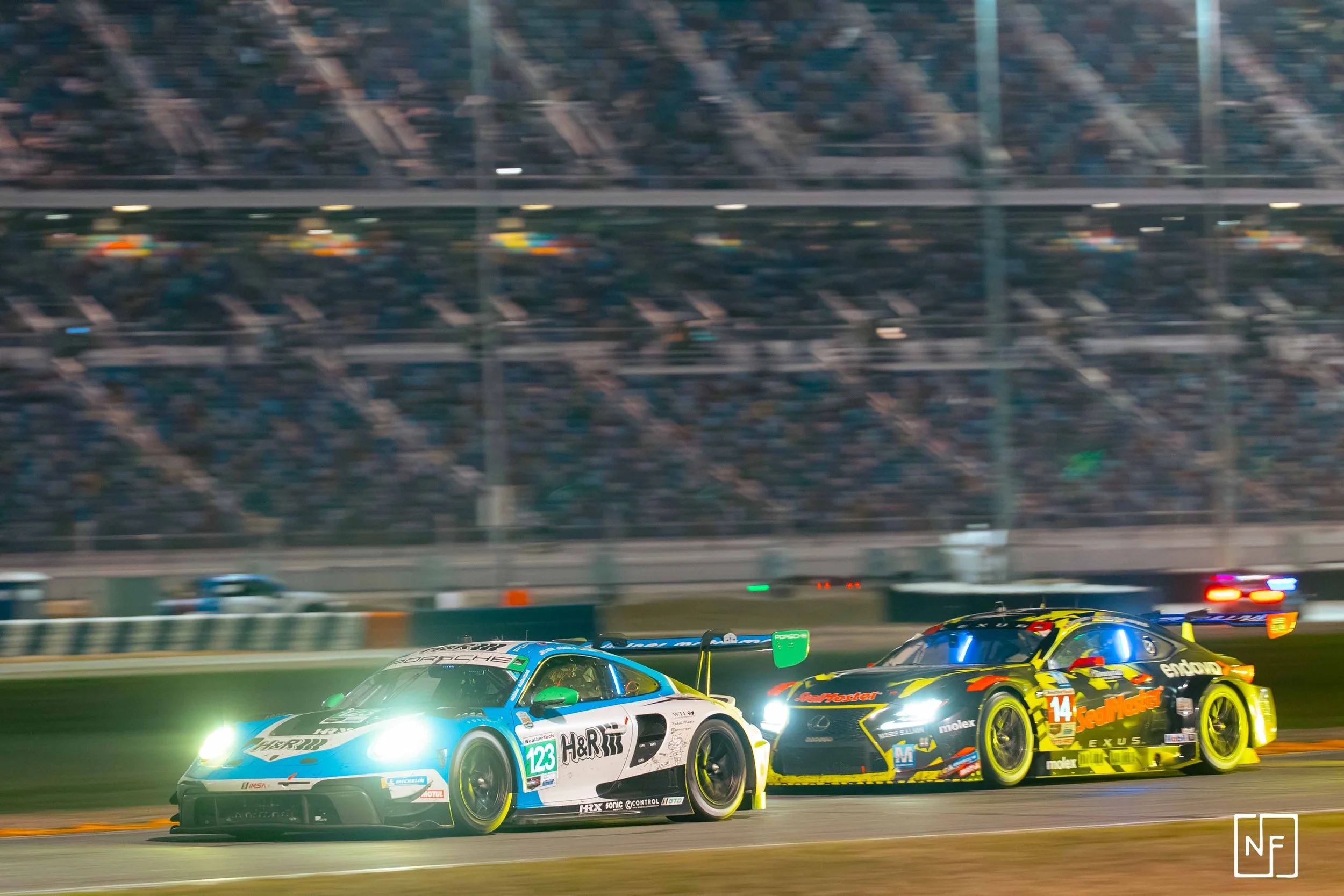 Two race cars competing on a track at night with a crowd in the background. One car is blue and white with the number 123, the other is black and yellow with the number 14. Bright headlights illuminate the scene.