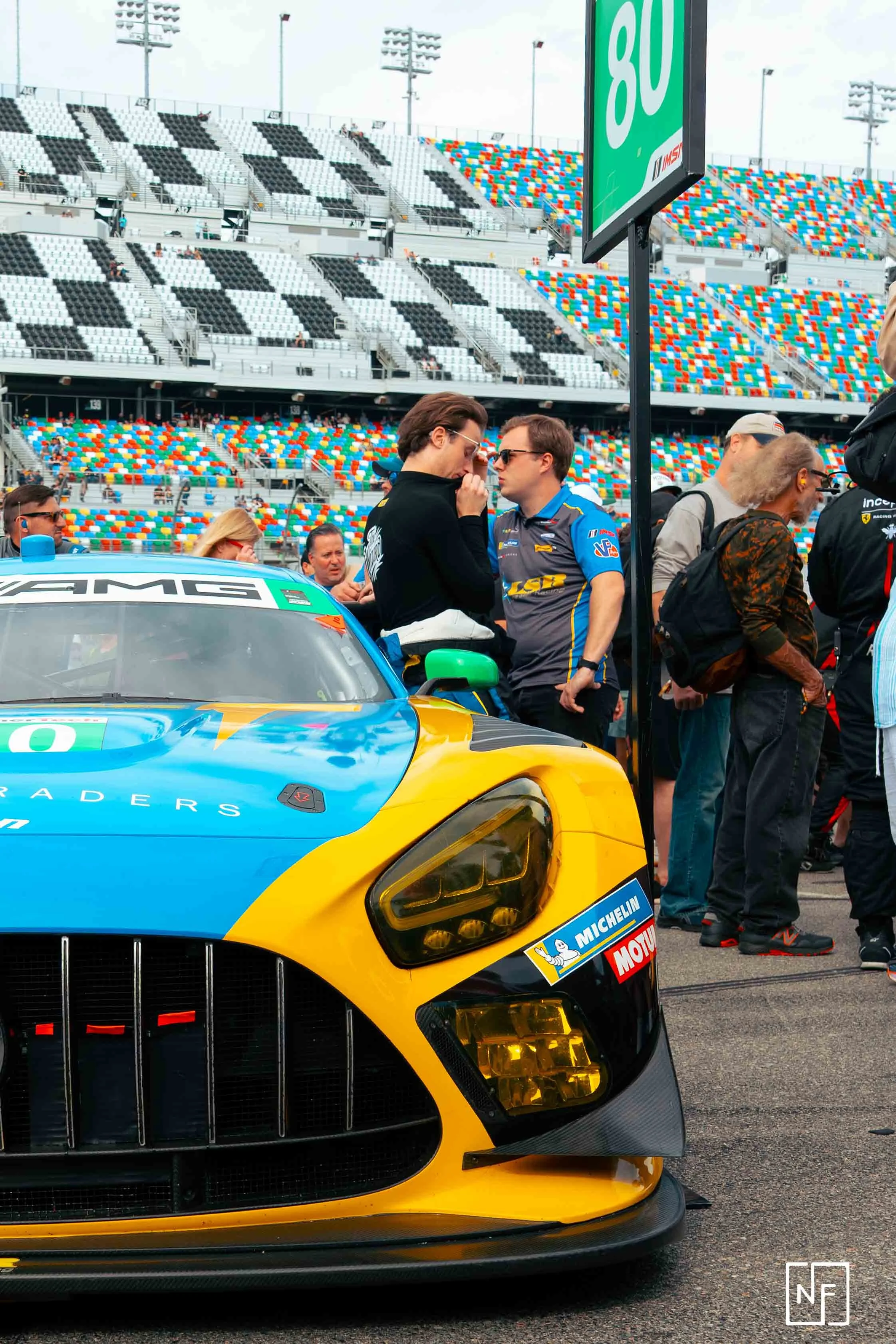 People gathered around a yellow and blue race car at a racetrack, with a large digital display showing '80' and empty grandstands in the background.