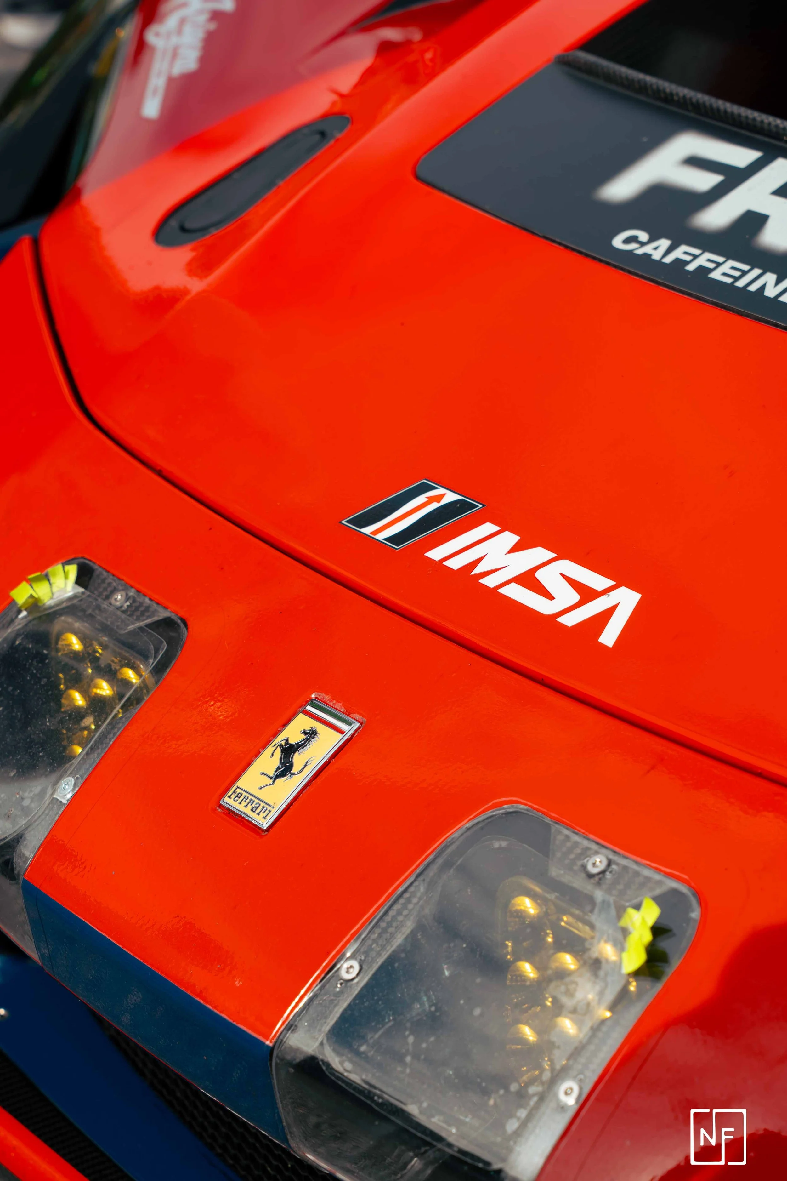 Close-up of a red Ferrari race car hood showing the Ferrari logo, IMSA sticker, and headlights with yellow fasteners.