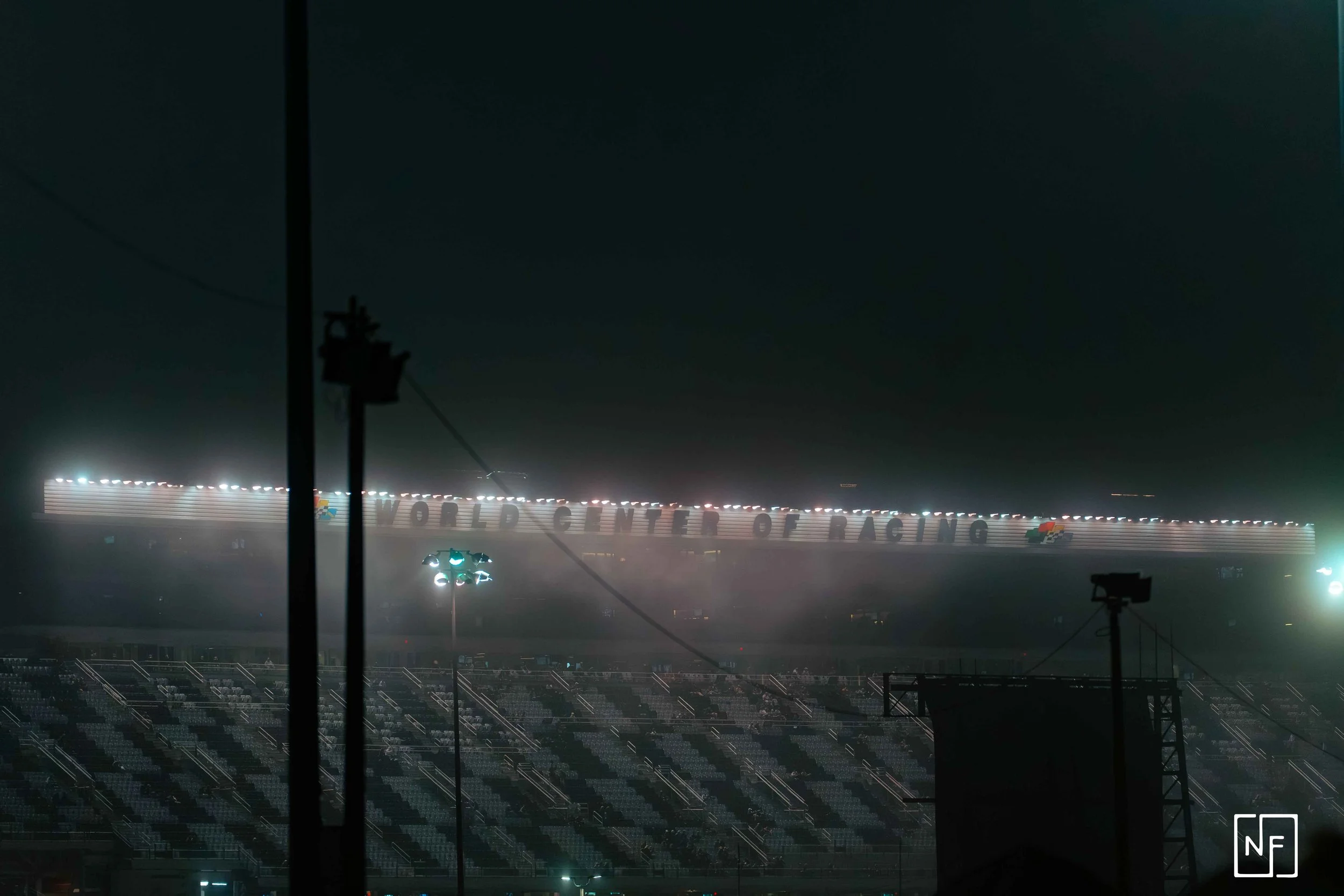 Nighttime view of the World Center of Racing sign on a grandstand at a racetrack, partially obscured by dark silhouettes and lighting fixtures.