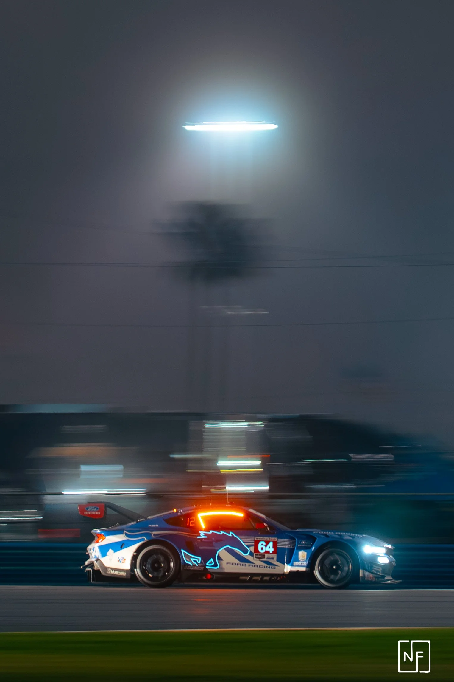 A racing car on a track at night, with motion blur and illuminated by bright lights, featuring the number 64 and a blue, white, and black design with a horse logo.