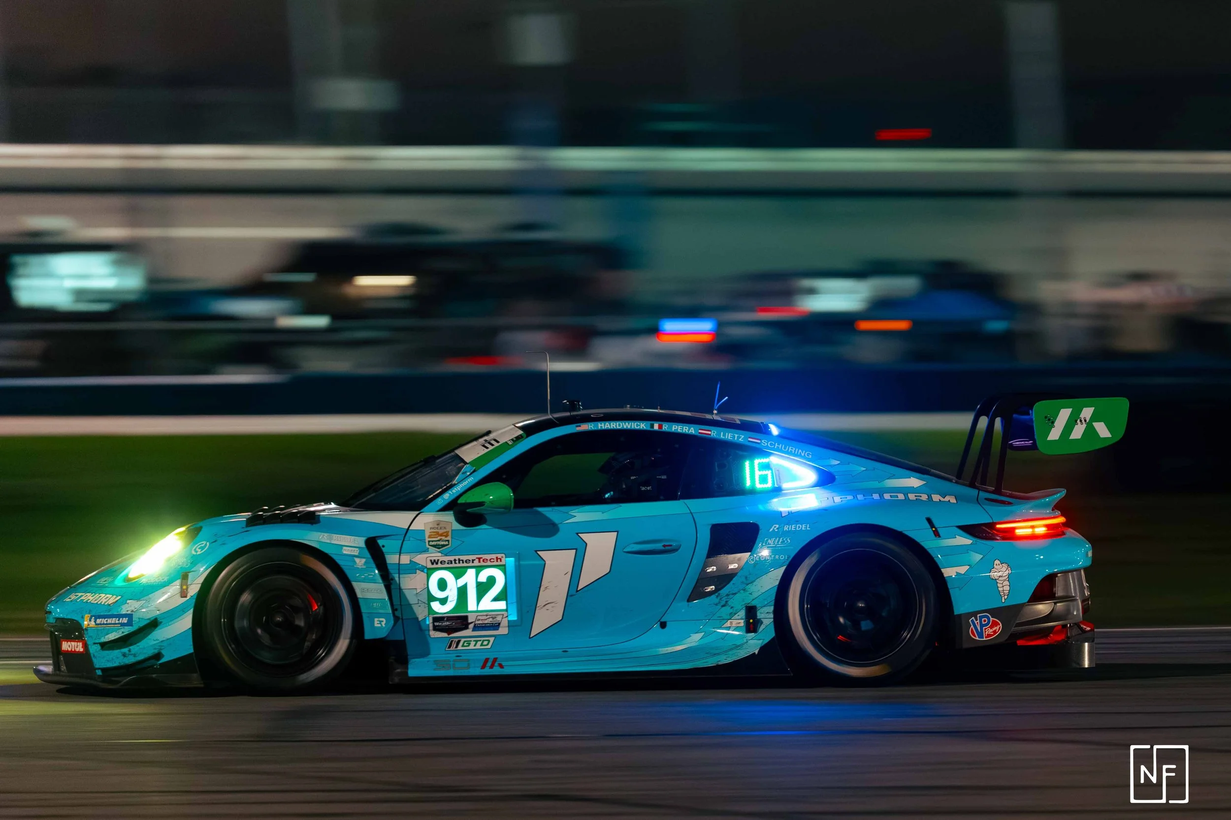 A blue race car moving at night on a race track, with blurred background and visible racing numbers 912 and 16 and various sponsor logos.