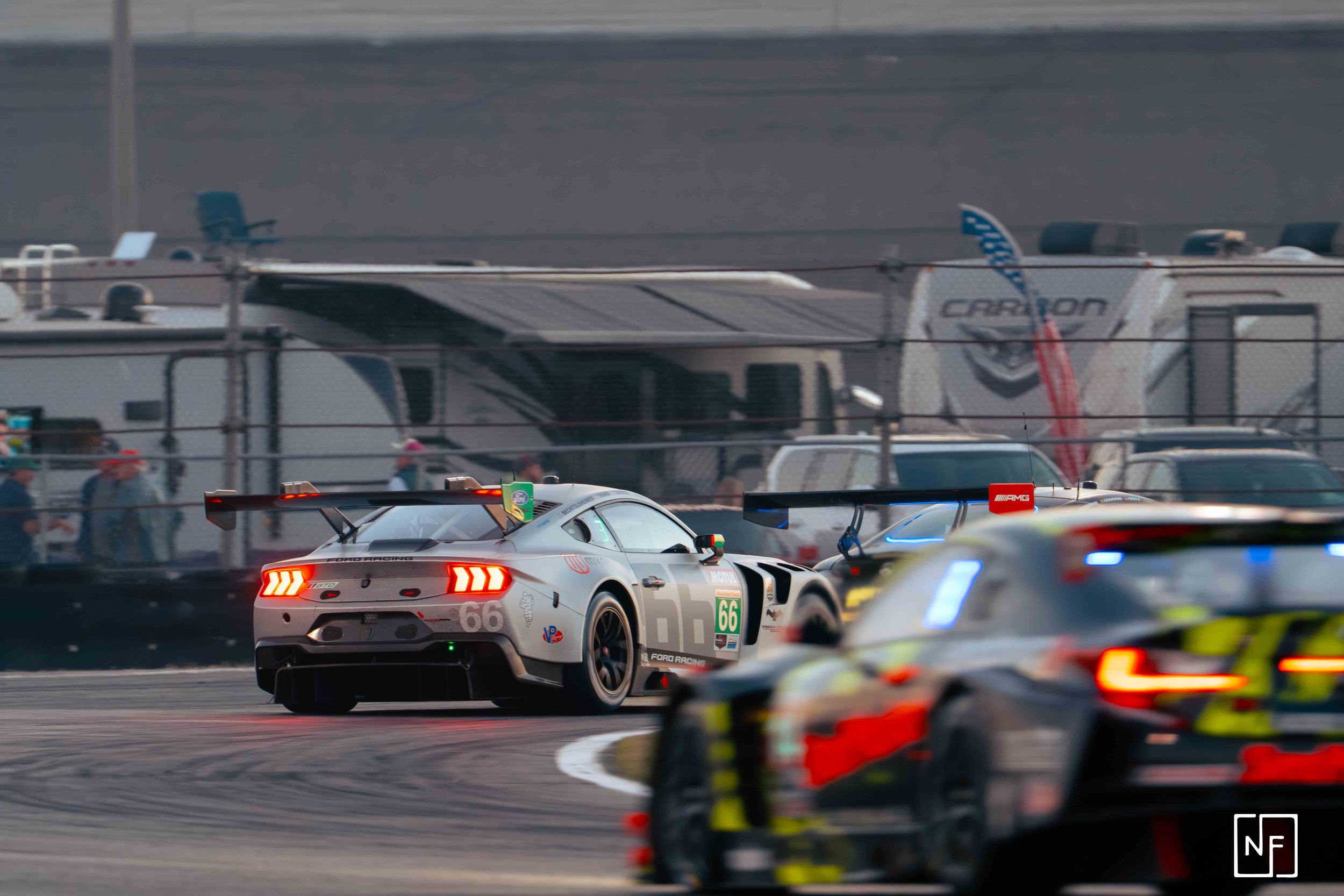 Race cars on a track during a motorsport event, with one car leading and others in pursuit, and trailers and spectators in the background.