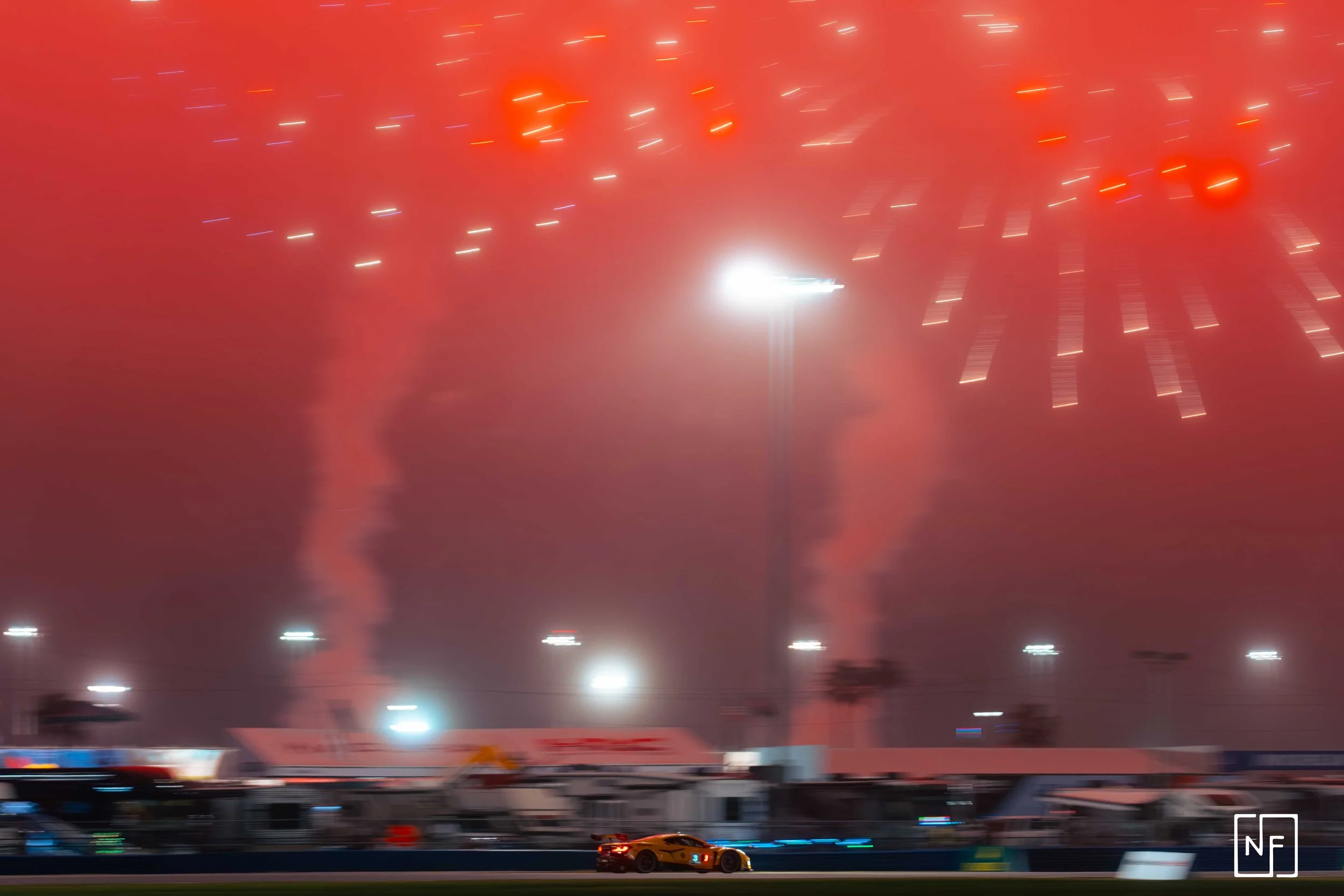 Nighttime fireworks display over a race track with racing cars and blurred background.