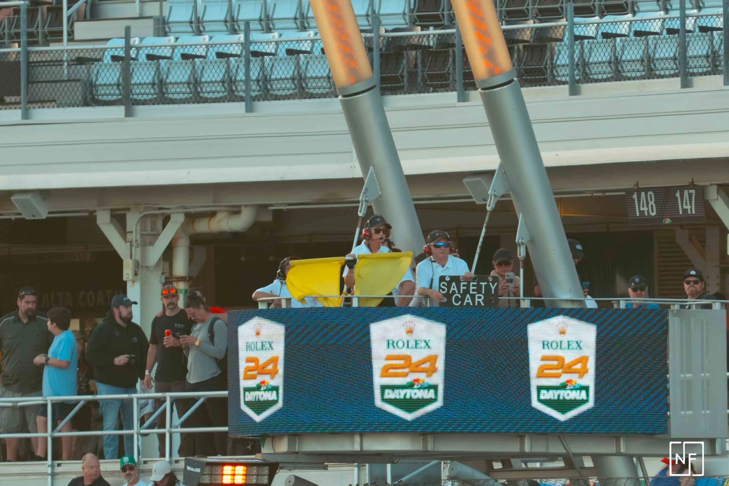 People in the monitoring area of a car race, with a large digital display showing the Rolex 24 at Daytona logo, and the people are wearing headphones and holding signs, including one that says 'SAFETY CAR'.