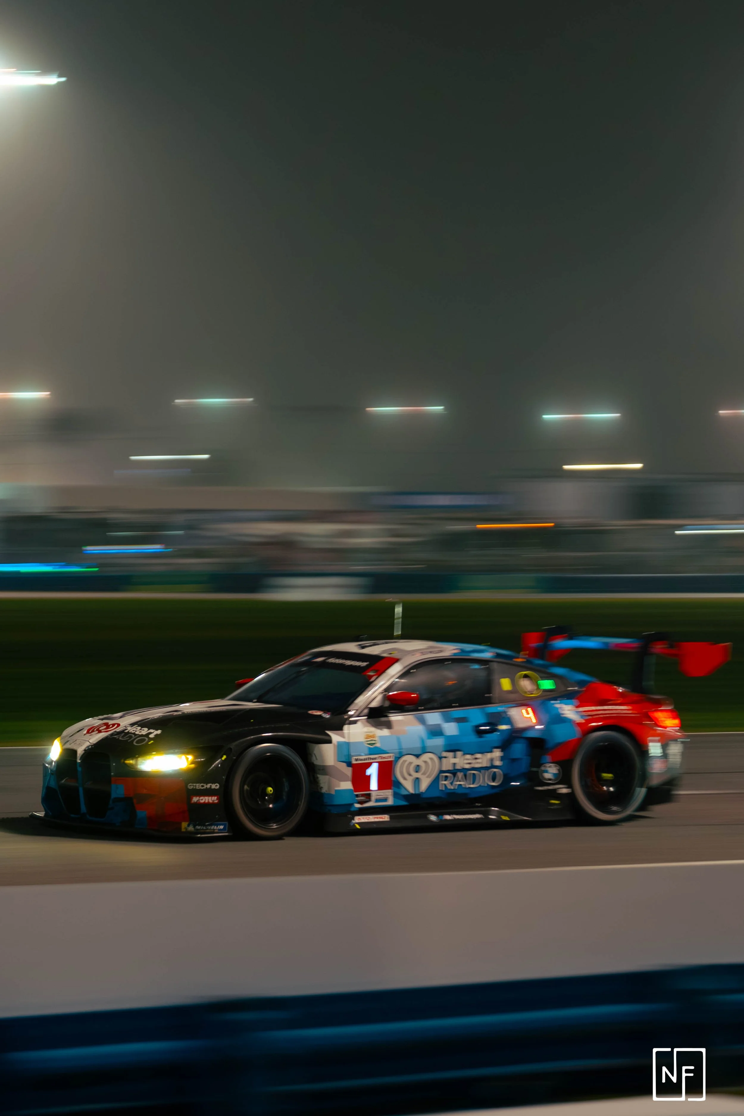 A racing car with a blue, black, white, and red livery speeding around a race track at night, with motion blur and bright headlights.