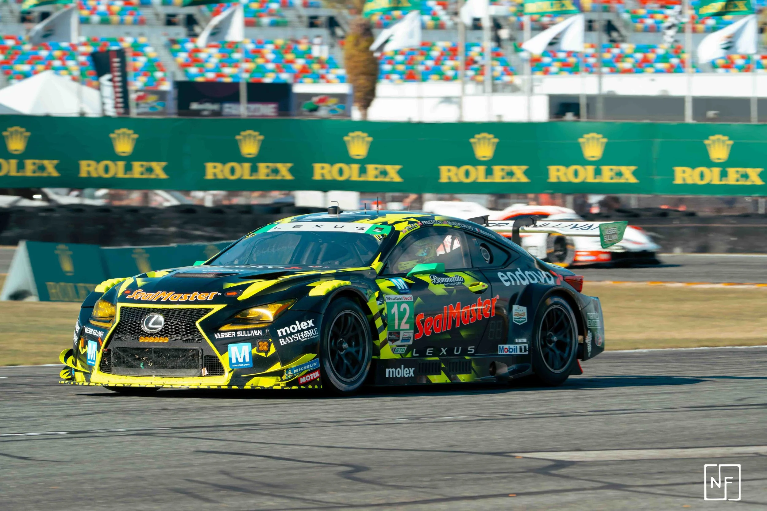 A black racing car with yellow accents speeding on a race track, with sponsors such as SealMaster, Lexus, and Motul visible. The track is lined with Rolex signage, and multiple cars can be seen in the background.