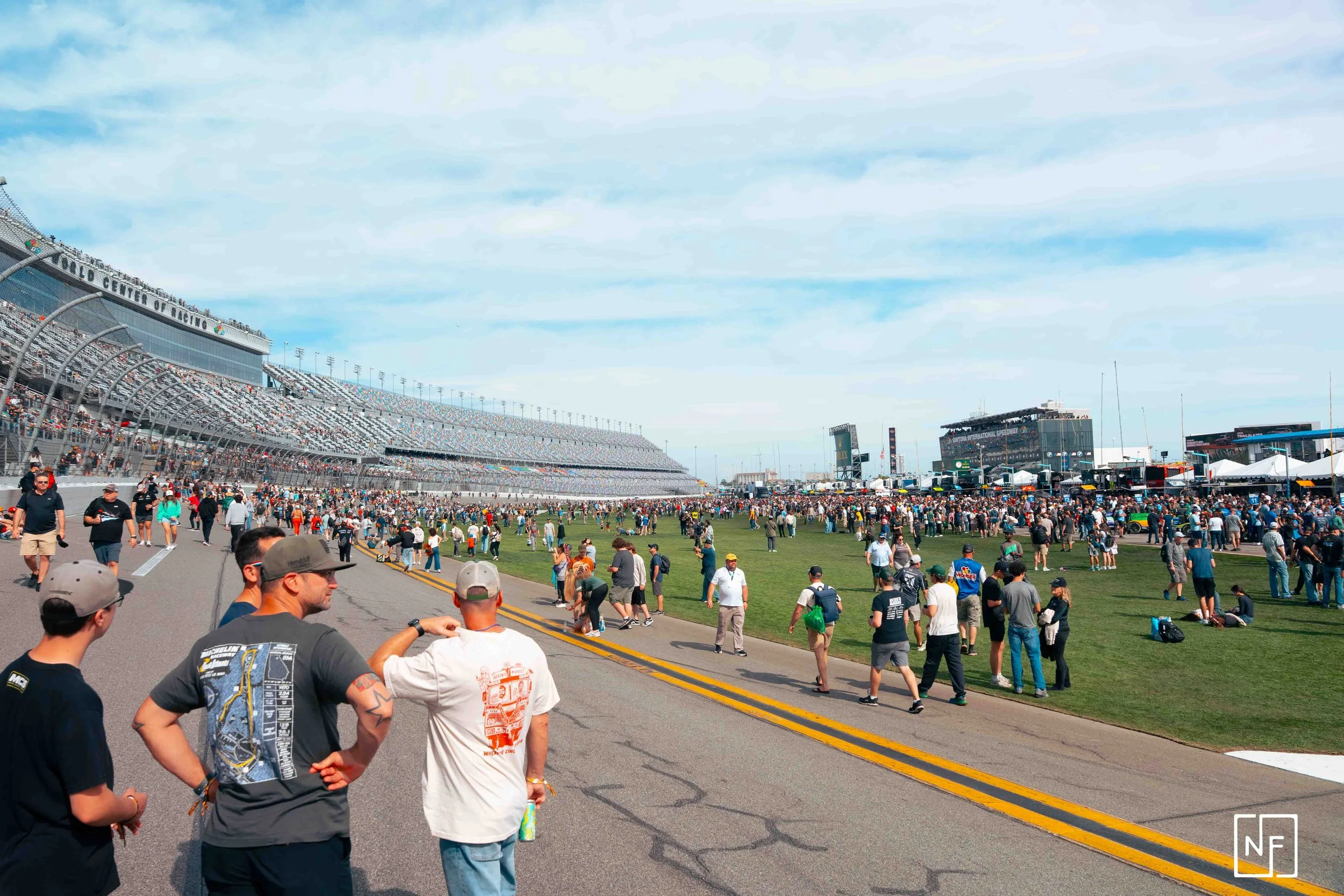 Crowd of people at a race track, with some walking on the paved road and others on the grass, large grandstands on the left and a stage with screens and tents on the right, under a partly cloudy sky.