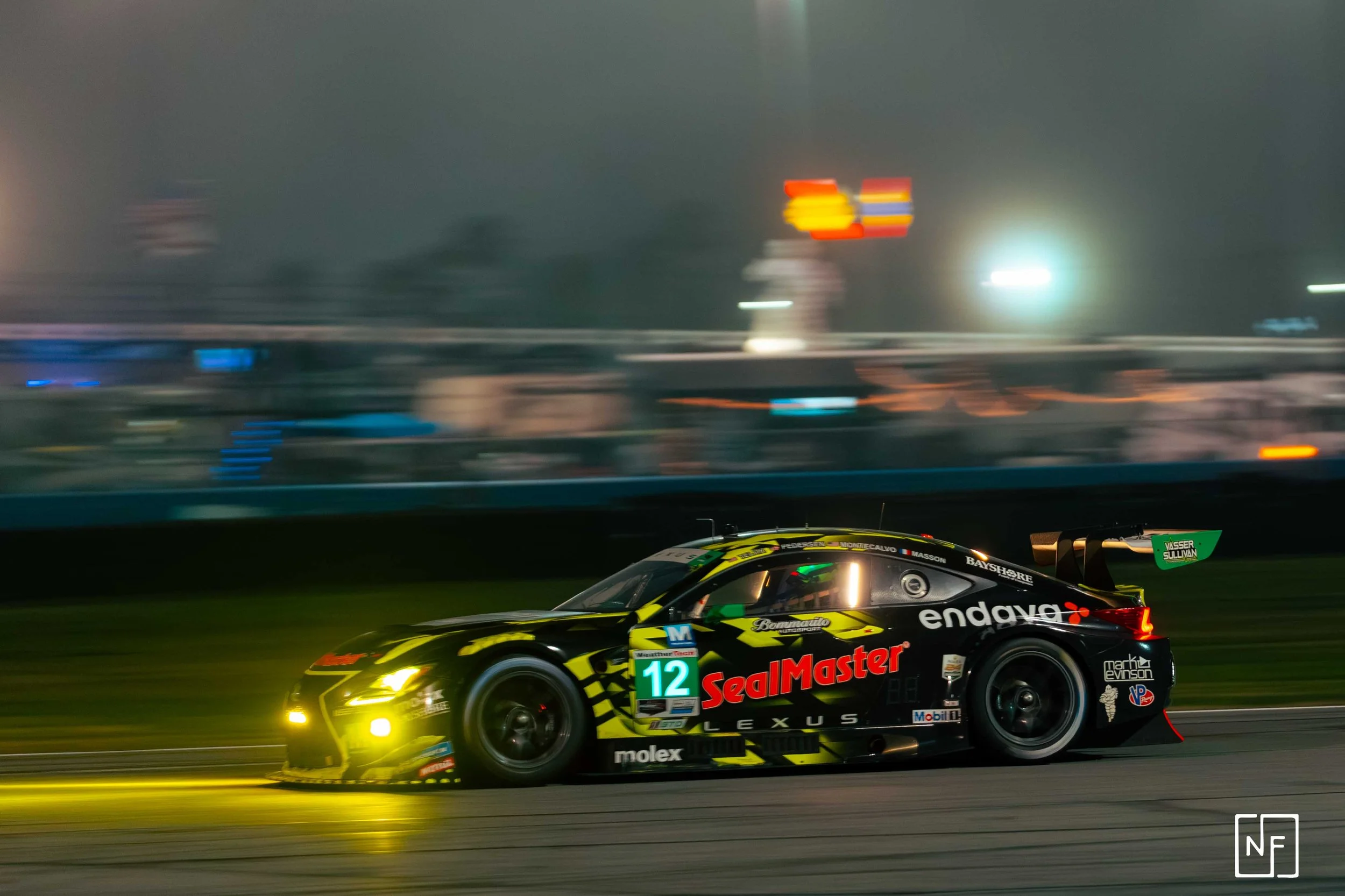 A black race car with yellow accents speeding on a racetrack at night, illuminated by headlights, with a blurred background of colorful lights and structures.