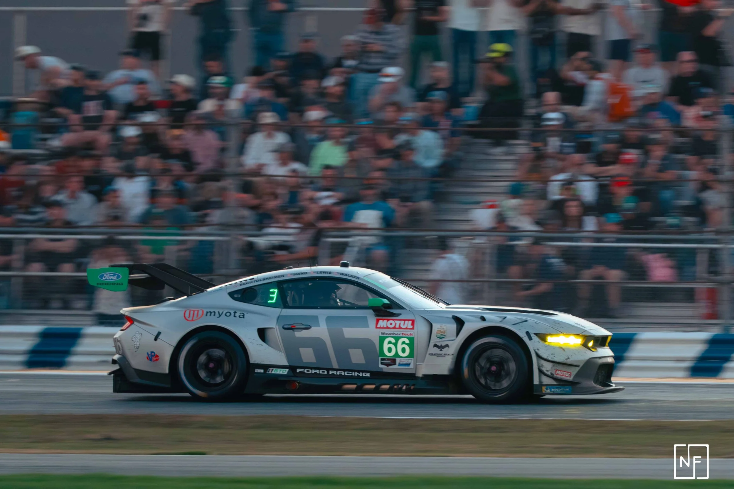 A white race car with the number 66 on its side speeds on a race track with a blurred crowd of spectators in the background.