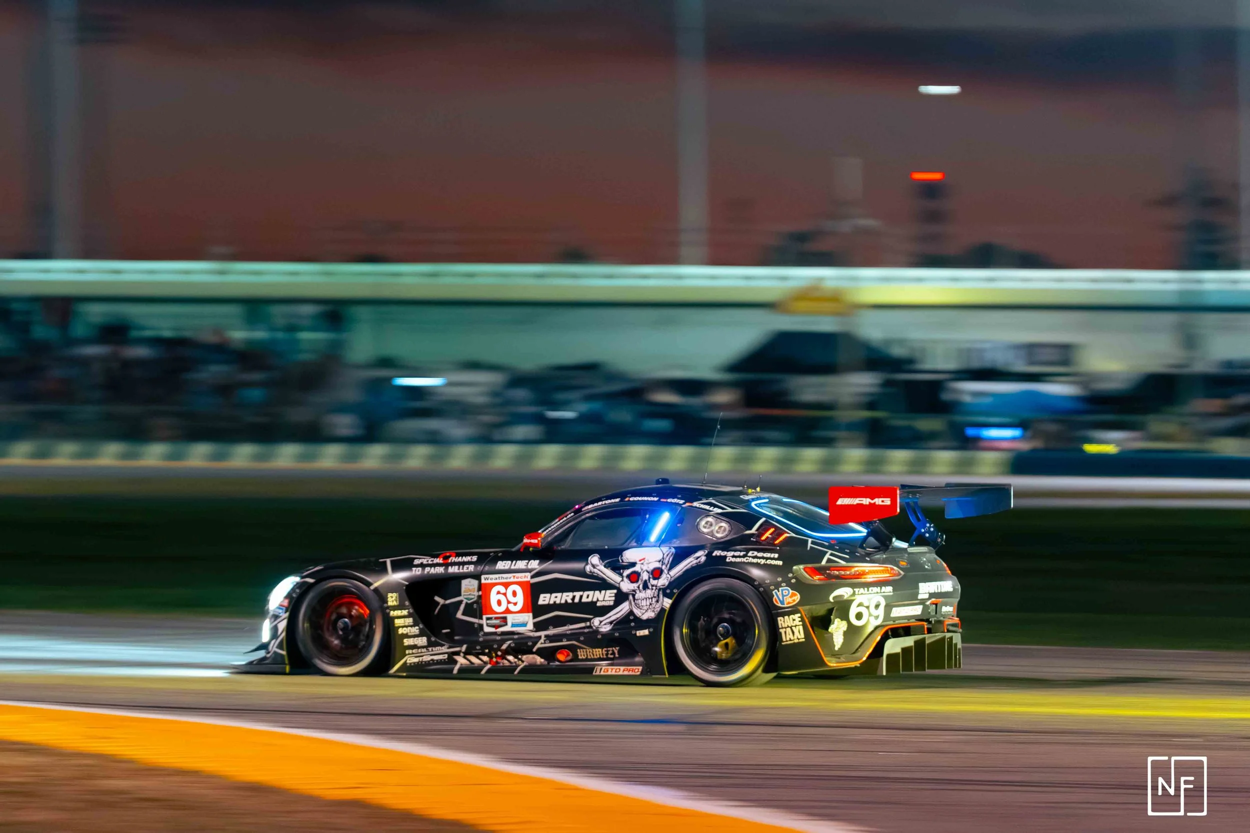 A black race car with the number 69, skull and crossbones graphic, and sponsor logos racing on a track at night.