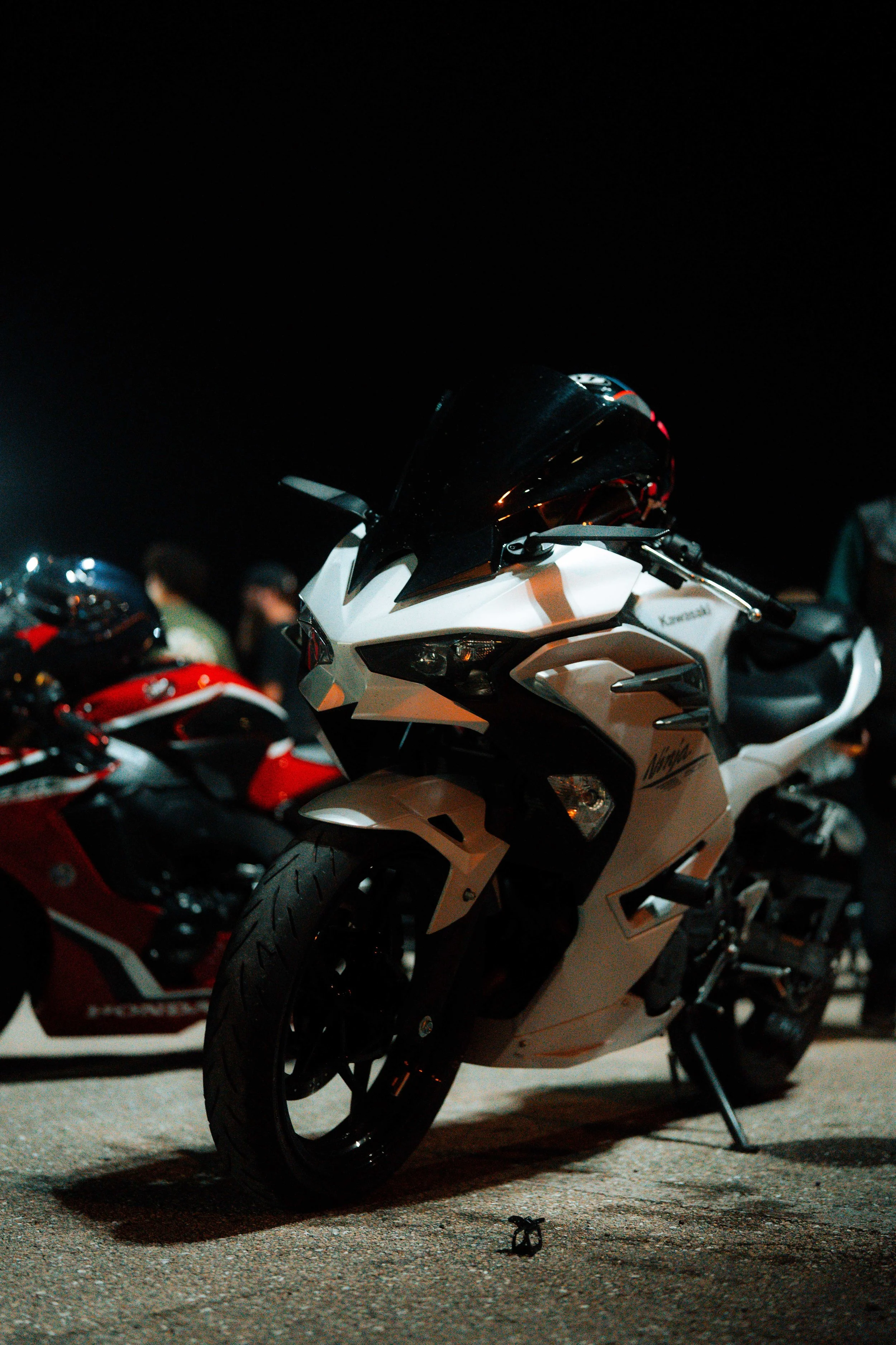 A white motorcycle parked on a street at night, with other motorcycles and people in the background.