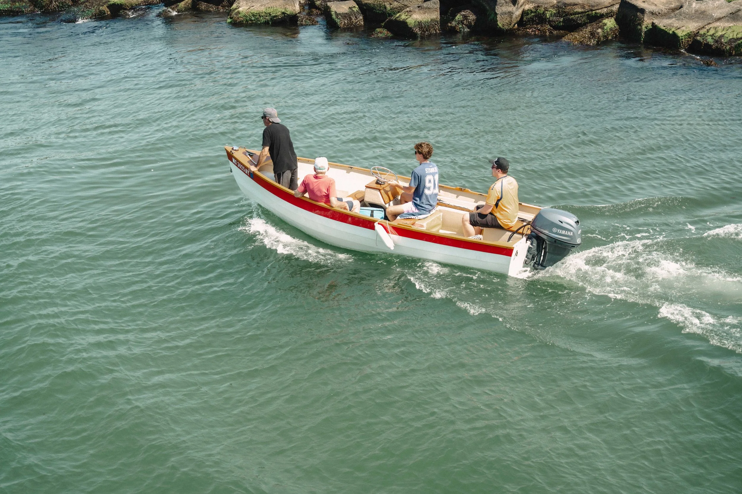 Four people in a small motorboat on a body of water, with rocky shoreline in the background.