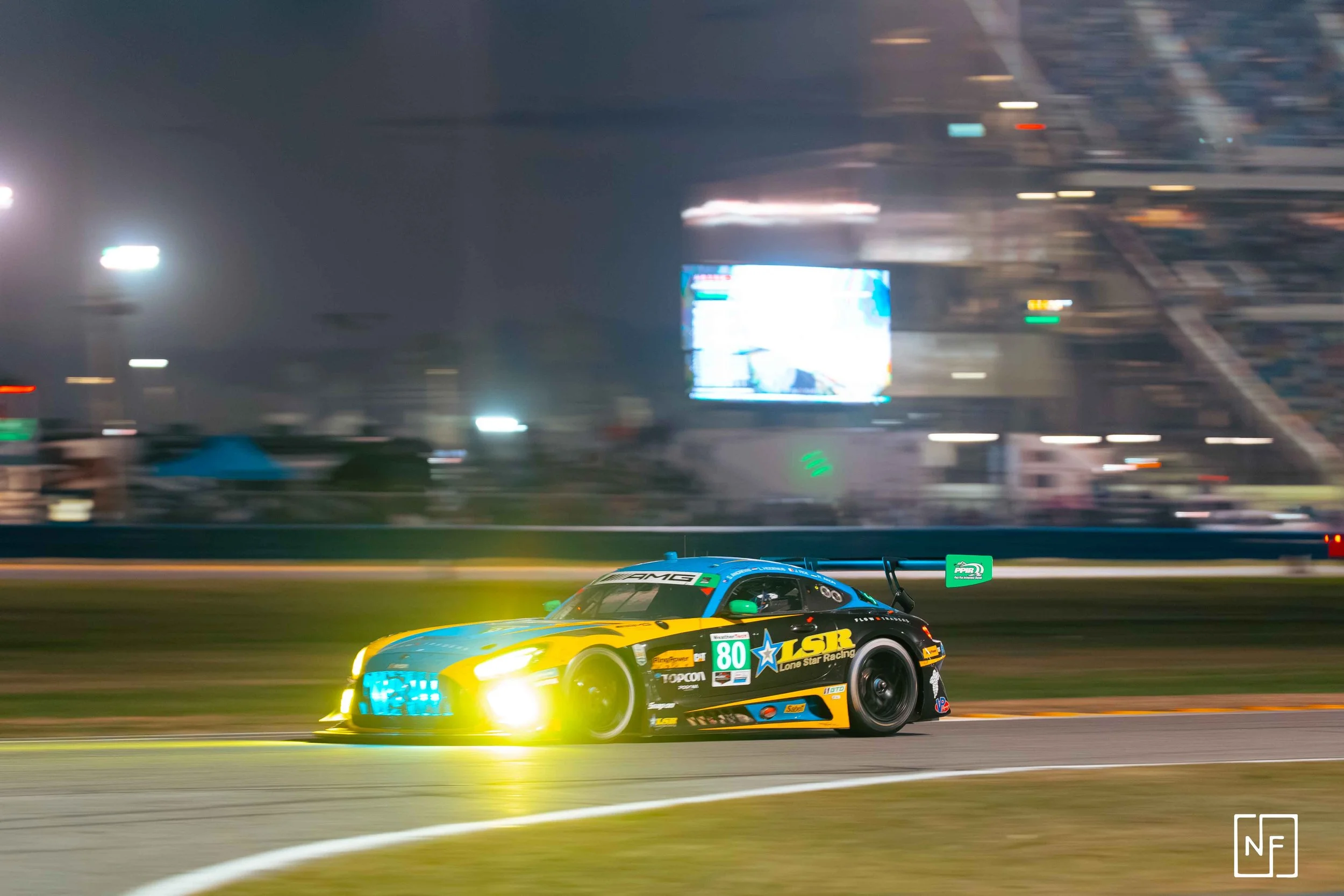 Nighttime race car on track with headlights on, brightly colored with blue, yellow, and black with sponsor decals, speeding past with blurred background of stadium and digital screens.
