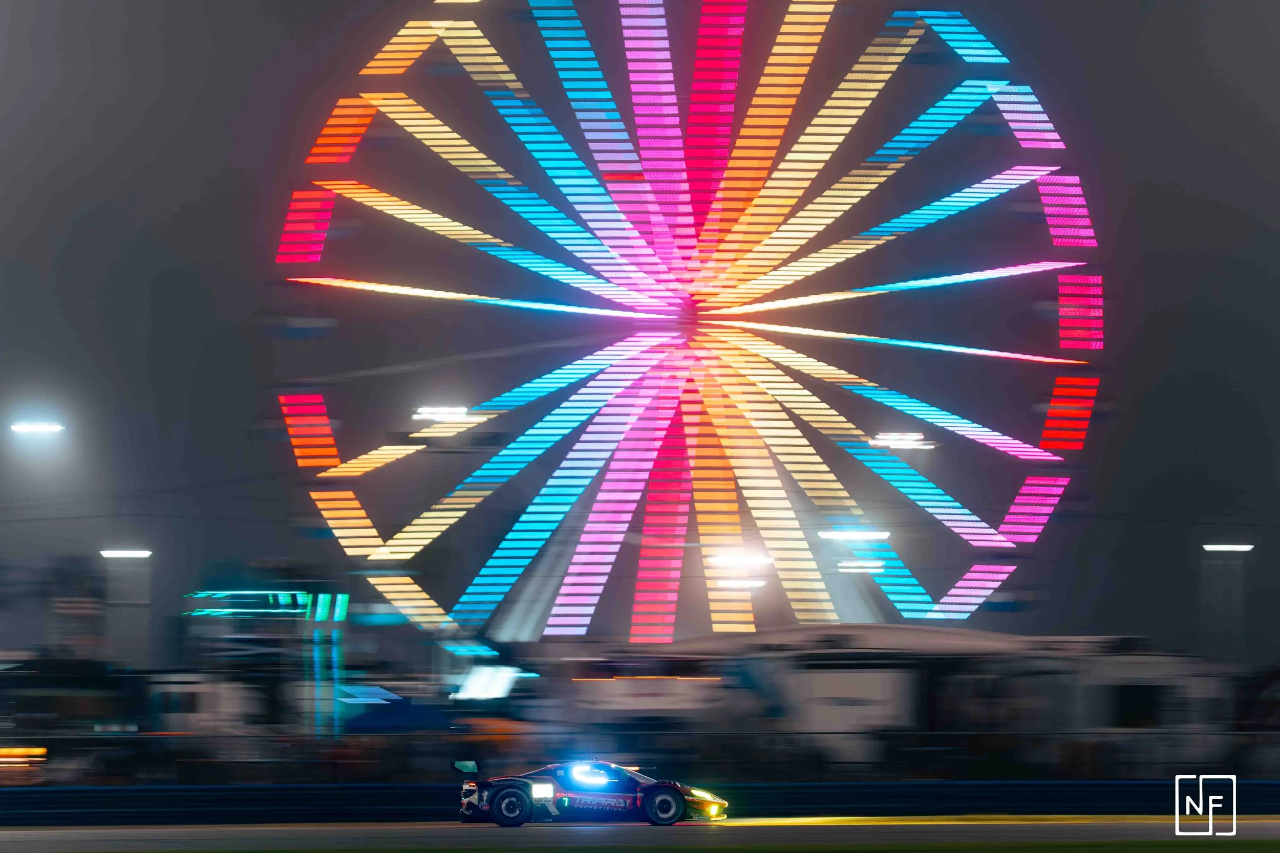 A race car speeds past a brightly lit carnival Ferris wheel at night. The Ferris wheel features colorful, illuminated sections creating a vibrant display against the dark sky.