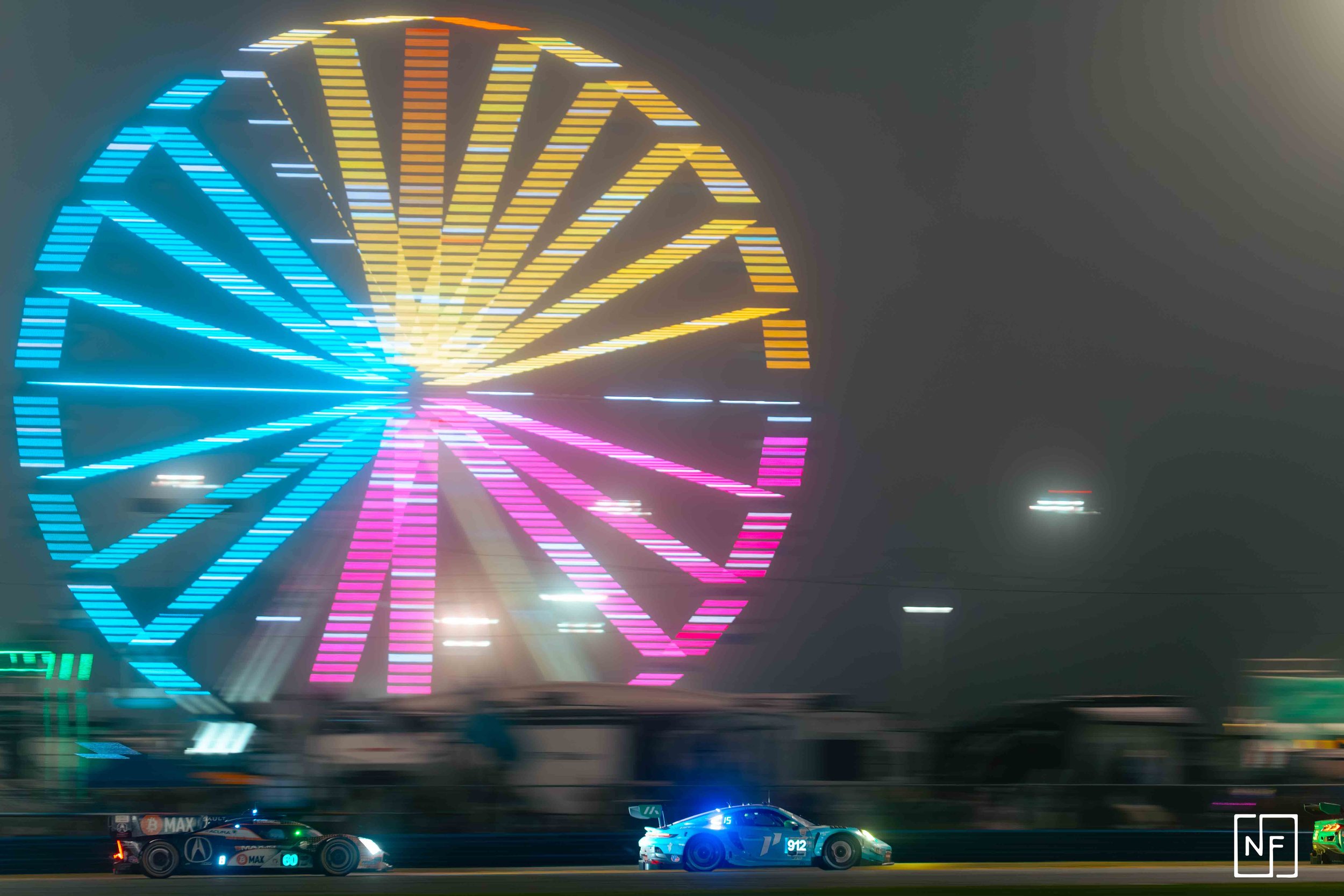Nighttime scene with brightly colored neon lights forming a large Ferris wheel in the background and two race cars speeding on a track in the foreground.
