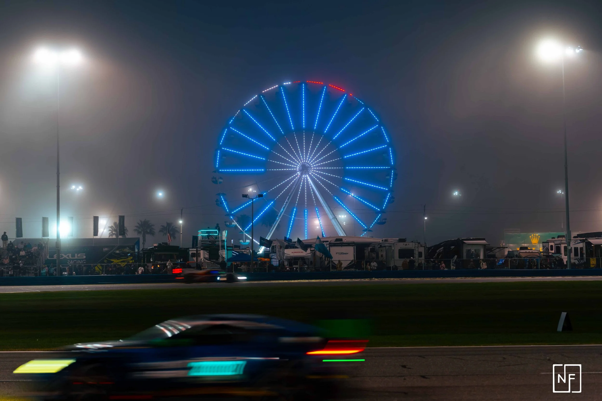 Nighttime scene at a fair with a brightly lit Ferris wheel in the background, a blurred race car passing on the track in the foreground, and foggy atmosphere with streetlights illuminating the area.