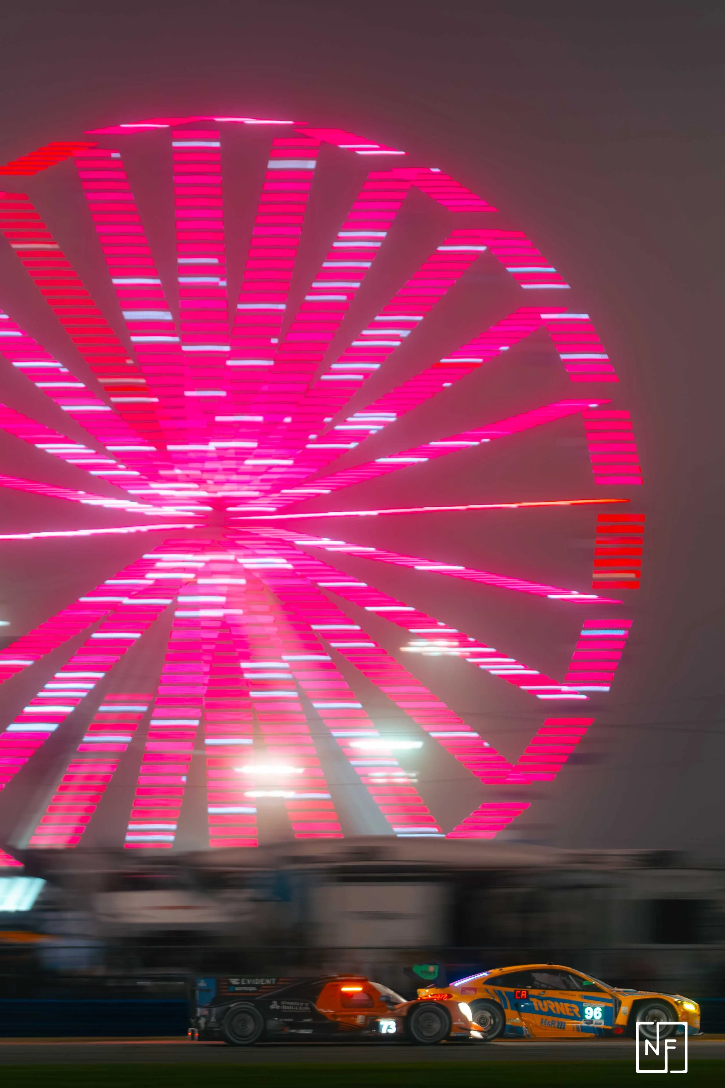 Two race cars speeding on a track at night with a large, pink-lit Ferris wheel in the background.