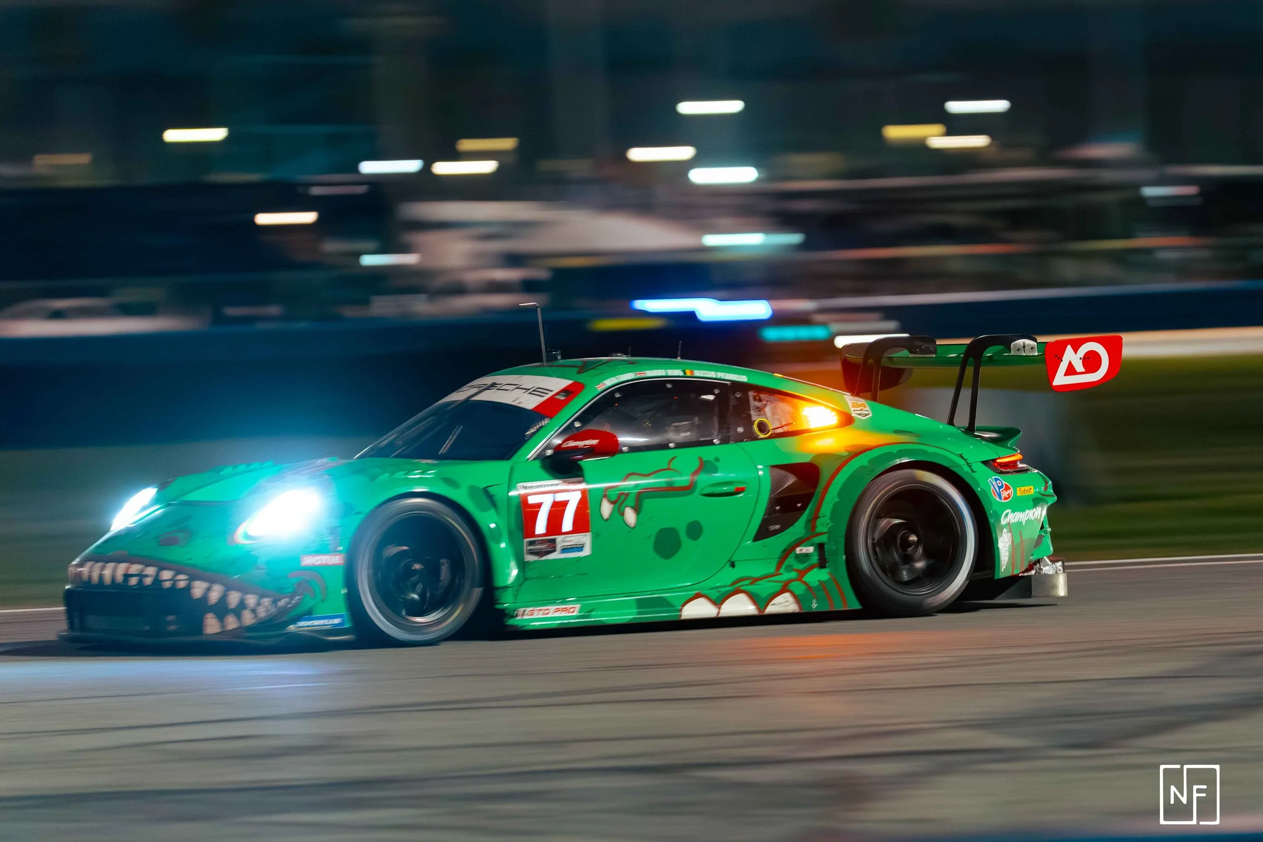 Green race car with number 77, designed to resemble a dragon or serpent, racing on a track at night with blurred background lights.